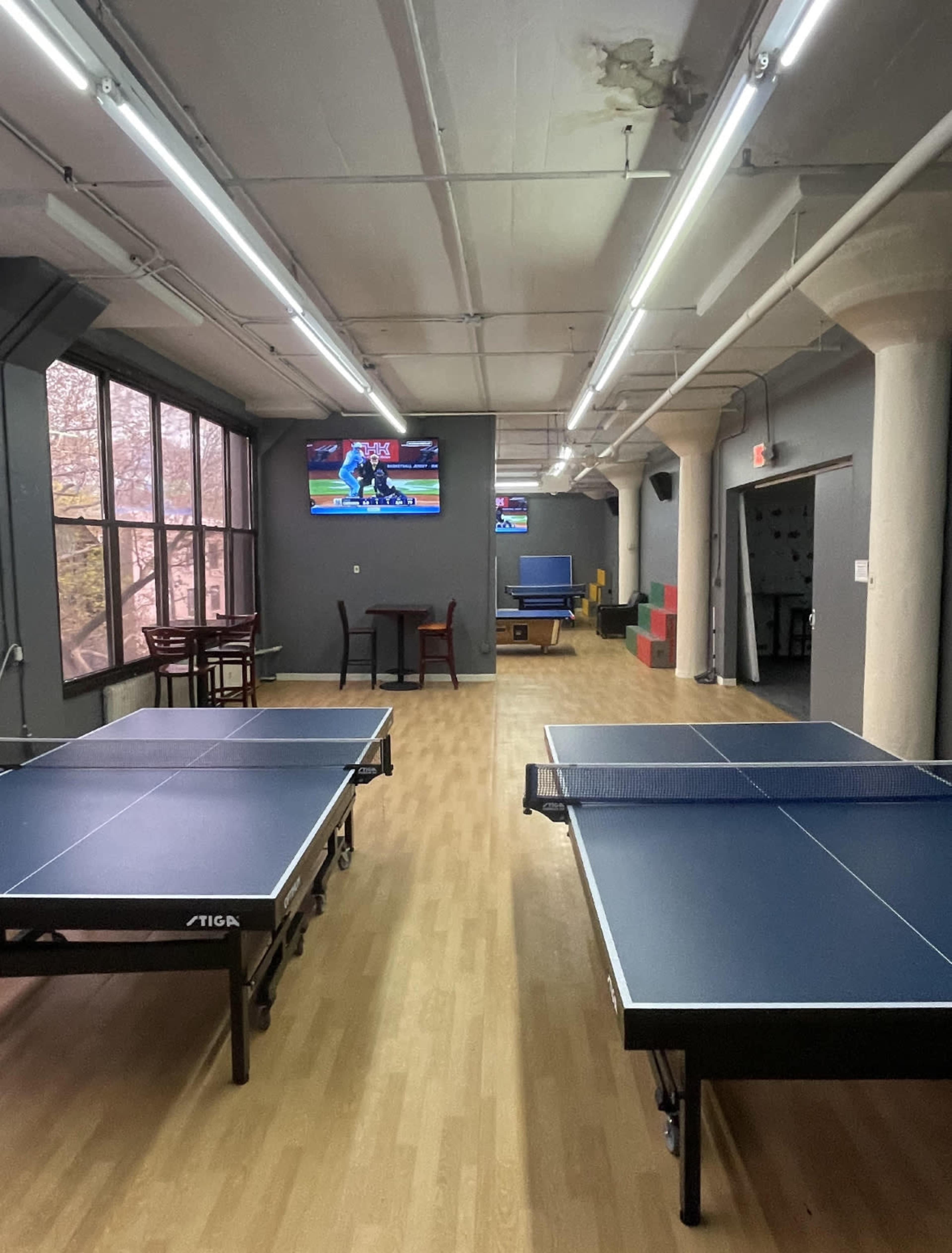 The image shows a spacious room with two table tennis tables, a seating area, and two televisions mounted on the walls.