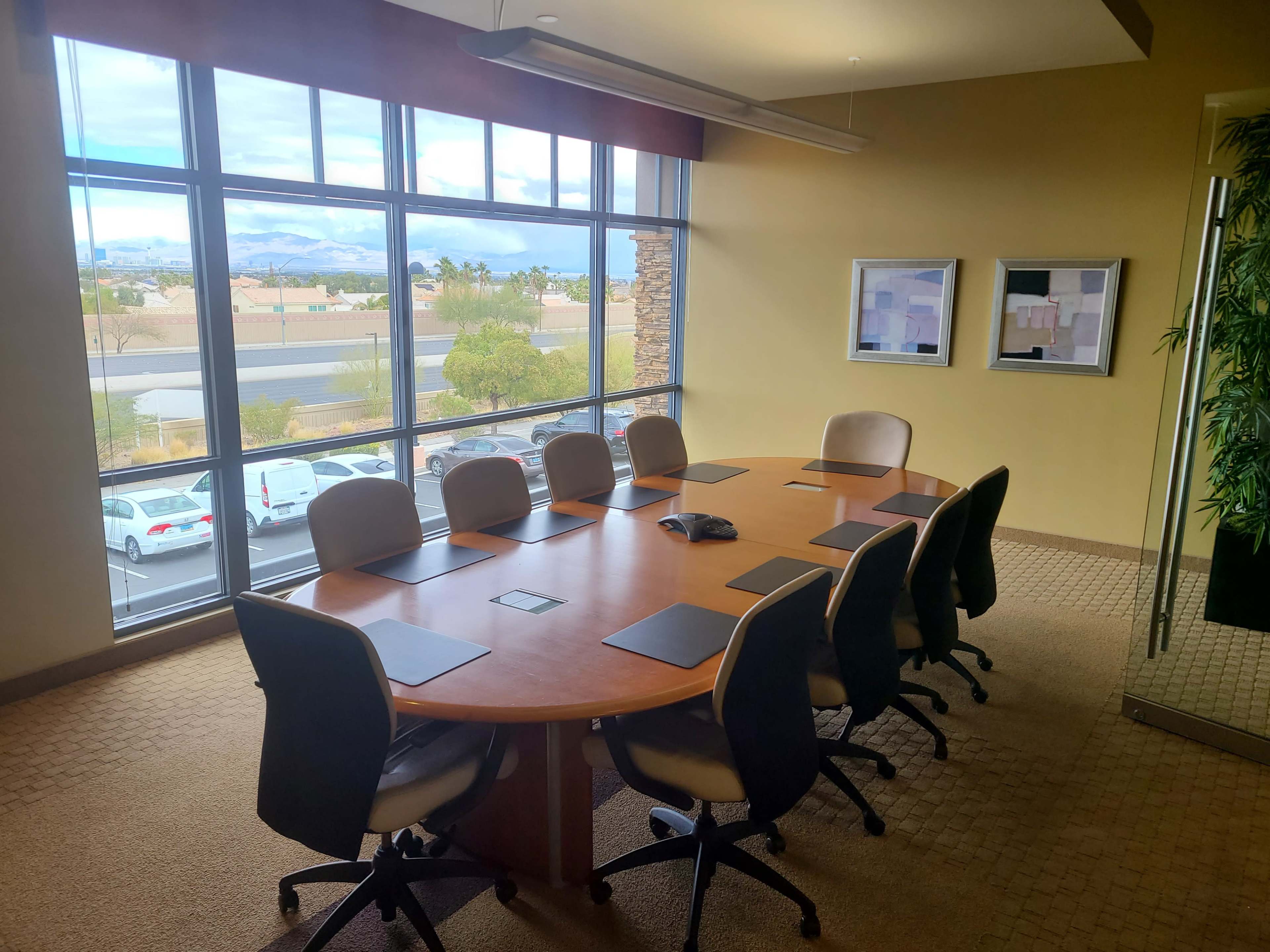 A conference room features a large oval wooden table surrounded by eight chairs, with large windows offering a view of the outside landscape.