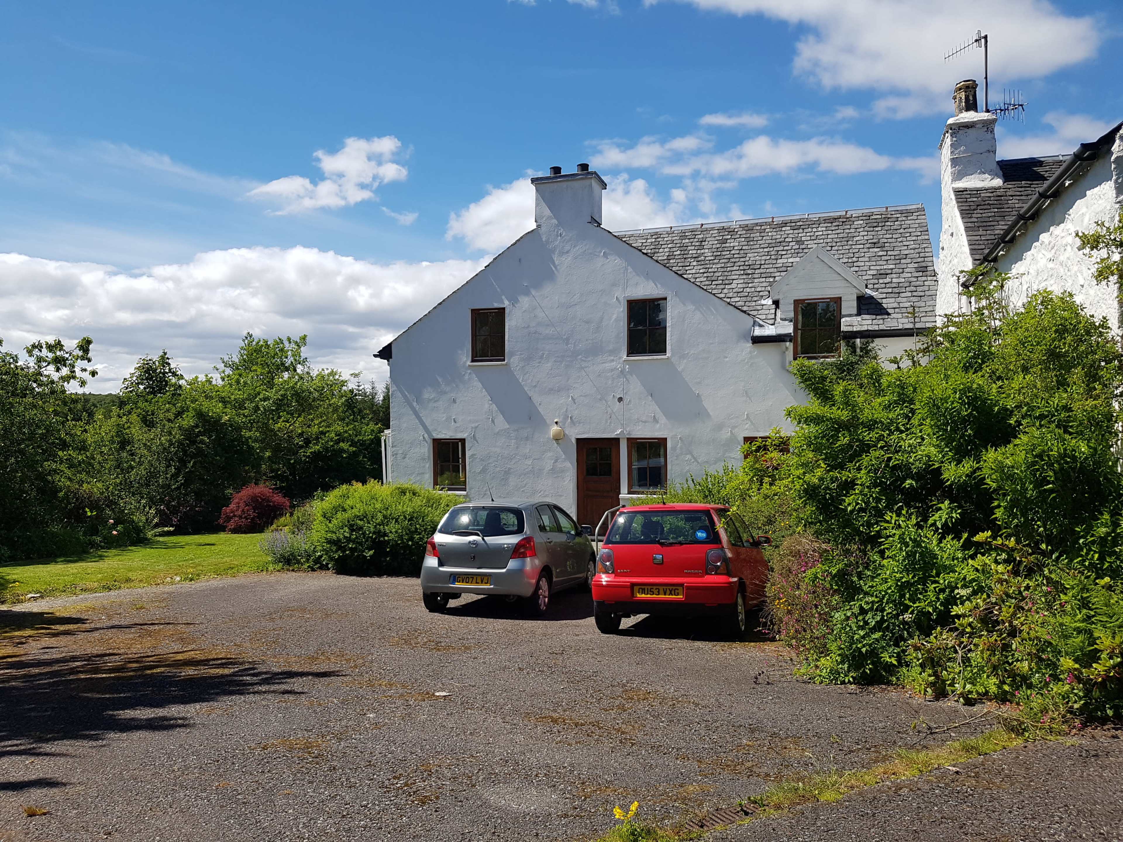 A white two-story house with a slate roof is situated next to a gravel parking area that contains two cars, surrounded by greenery and under a partly cloudy sky.