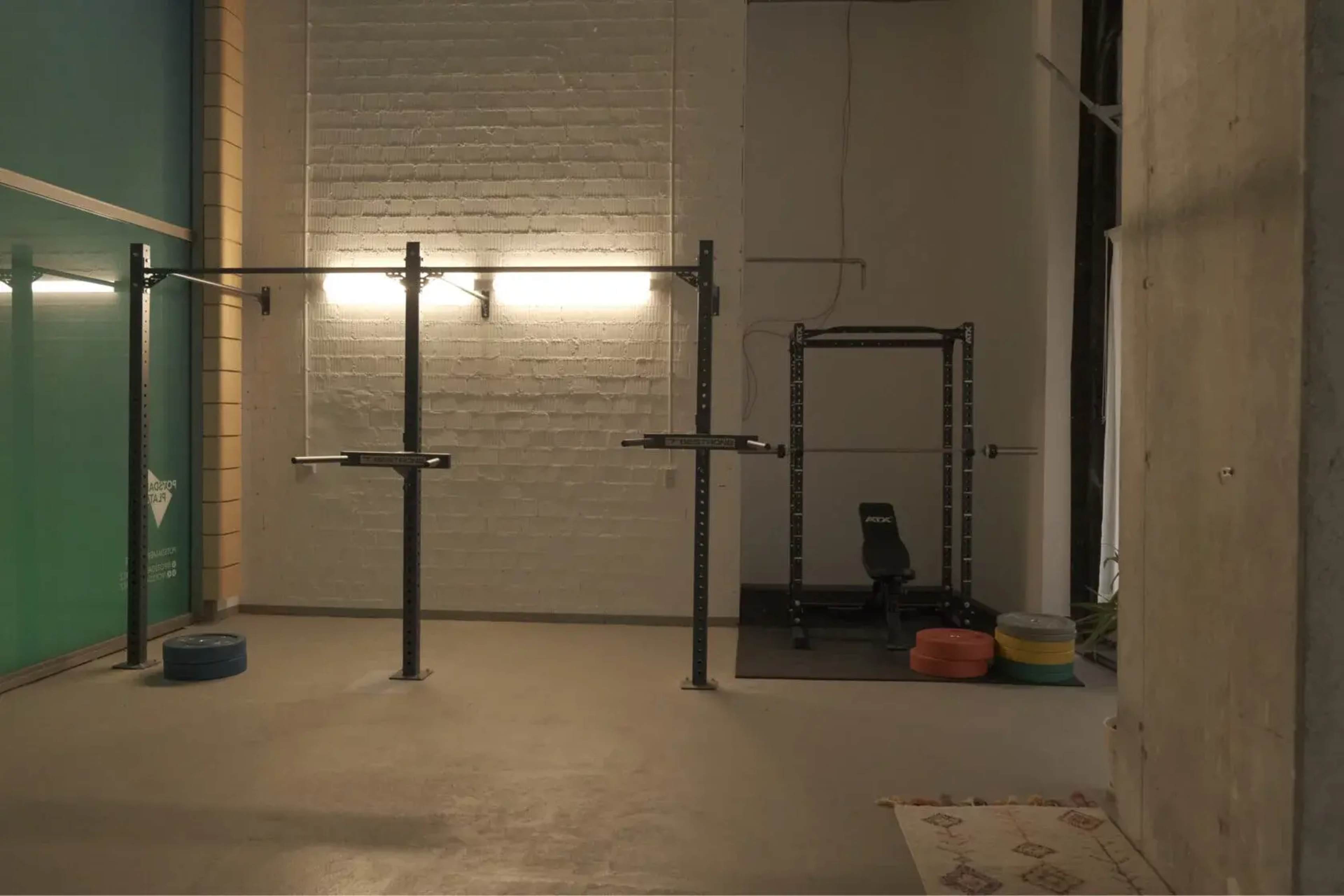 The image shows a spacious gym area featuring pull-up bars, a squat rack, and weight plates arranged on the floor.