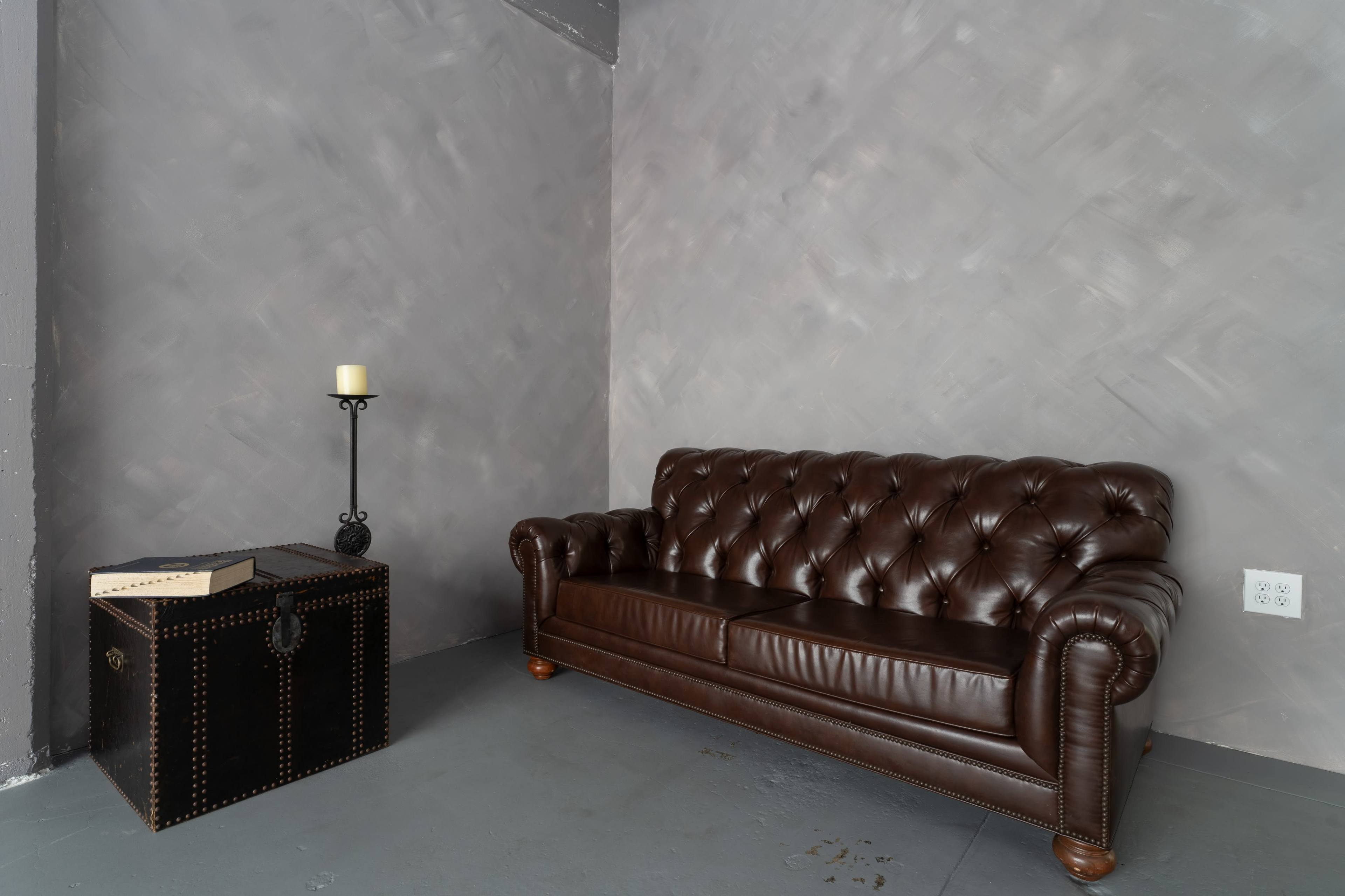 A brown leather chesterfield sofa and a black trunk with a candle holder are positioned against a gray wall.