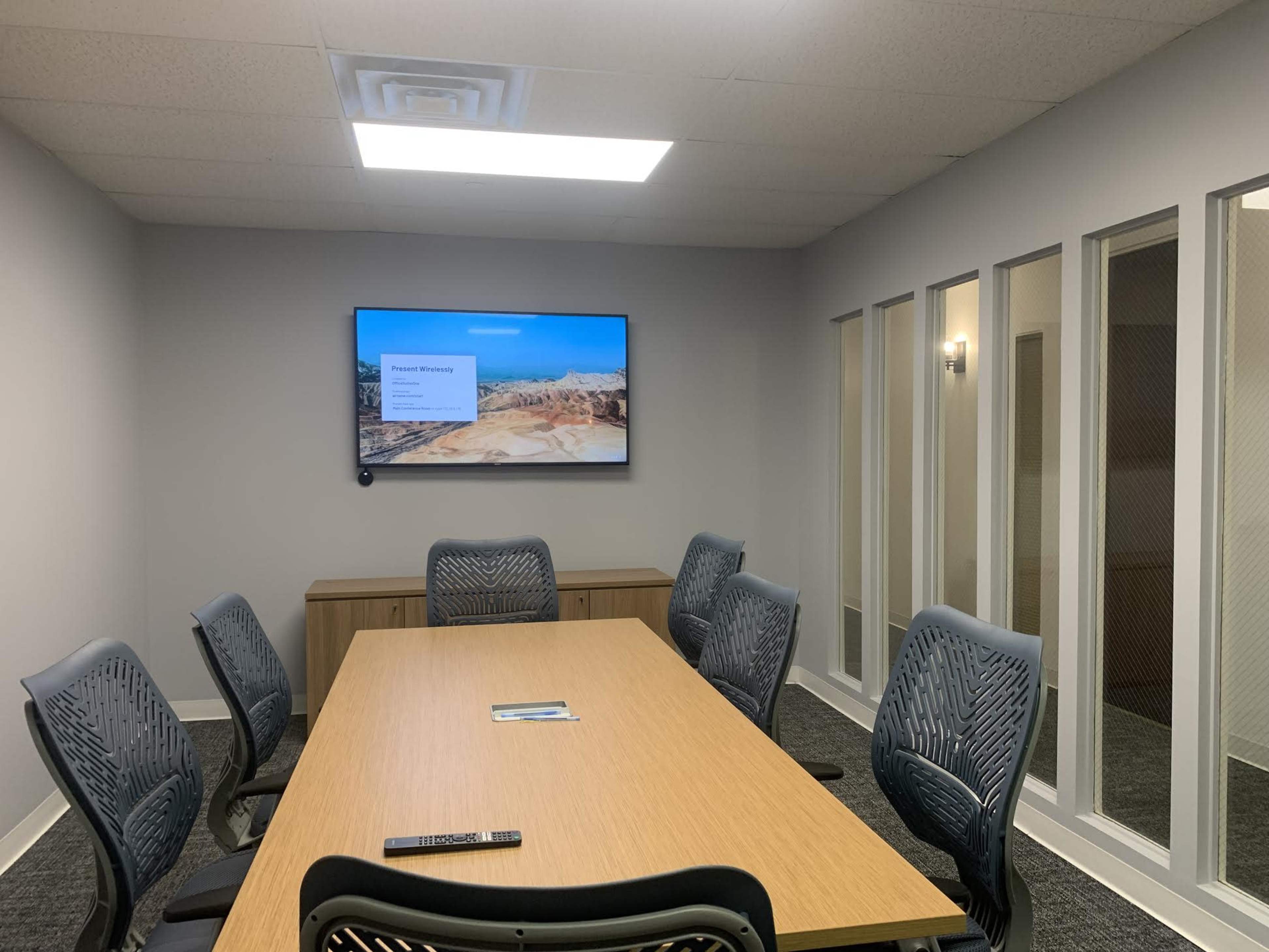 A conference room features a large wooden table surrounded by ergonomic chairs, with a mounted screen displaying a presentation and glass-paneled walls separating it from an adjacent space.