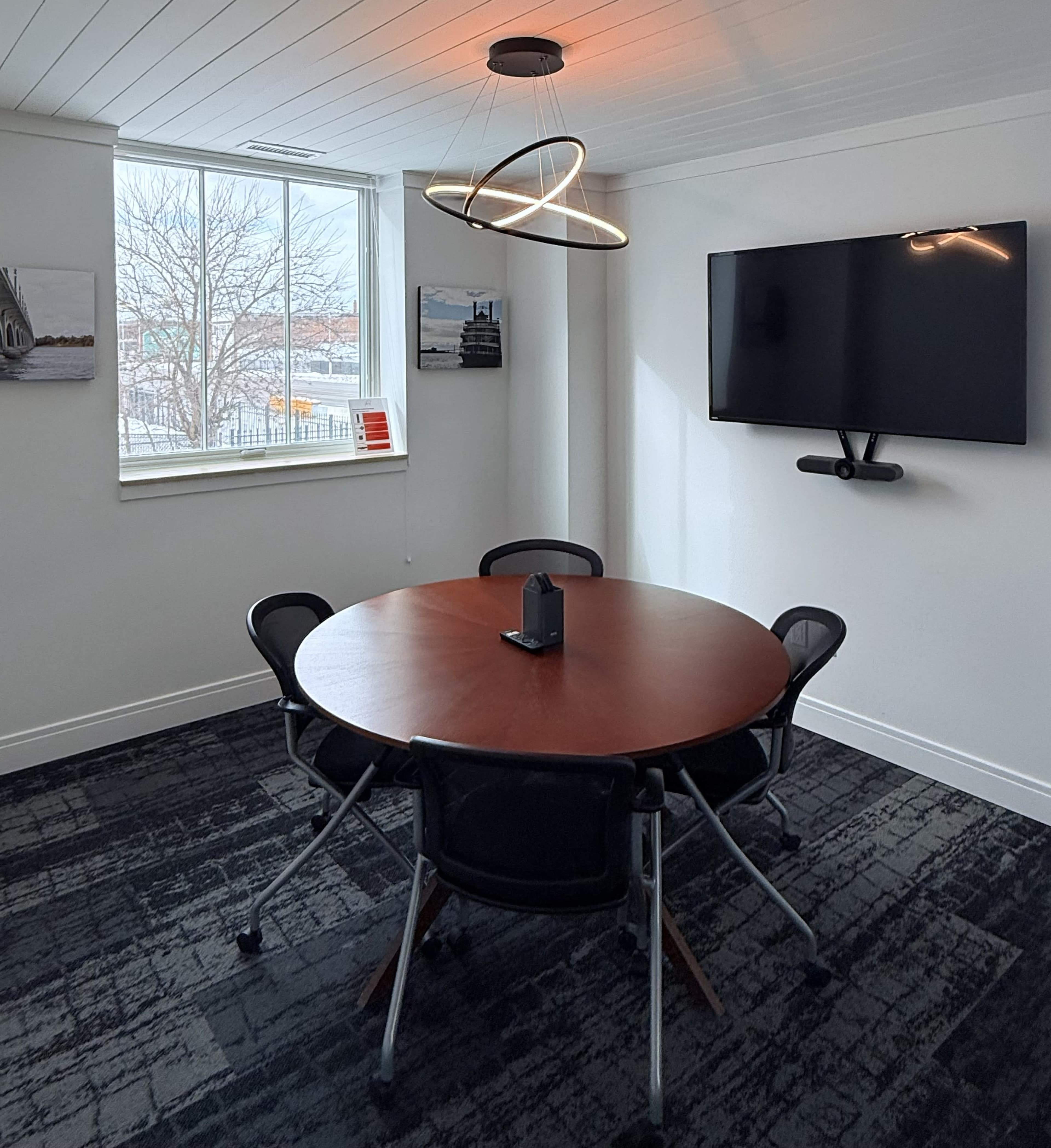 The image shows a small conference room with a circular wooden table, six black chairs, a television mounted on the wall, and two framed pictures near a large window.