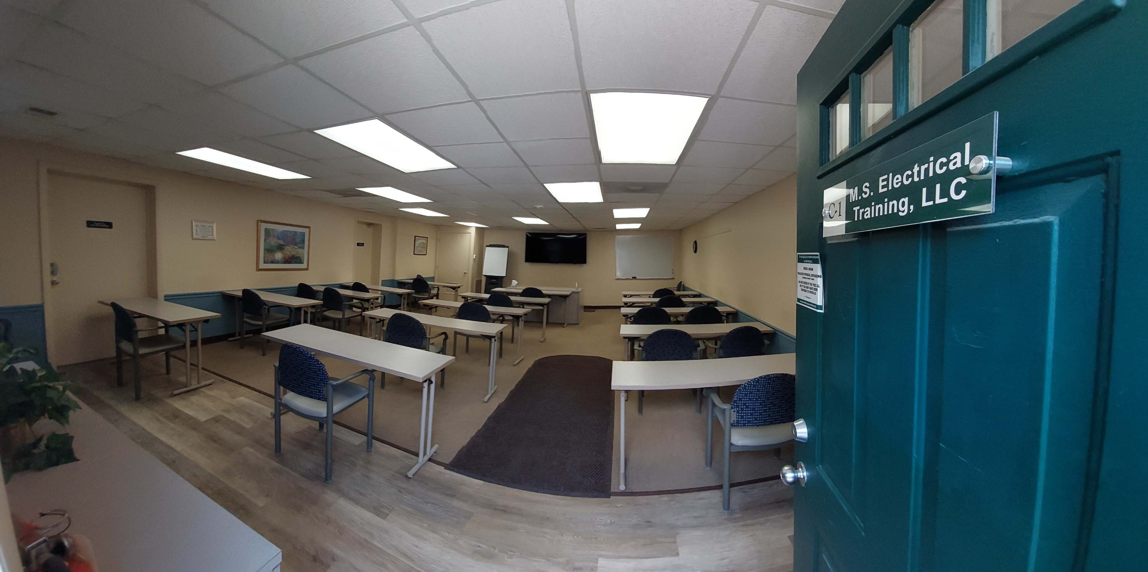 A spacious training room with multiple tables and chairs arranged in neat rows, featuring a large screen at one end and natural light entering through overhead lights.