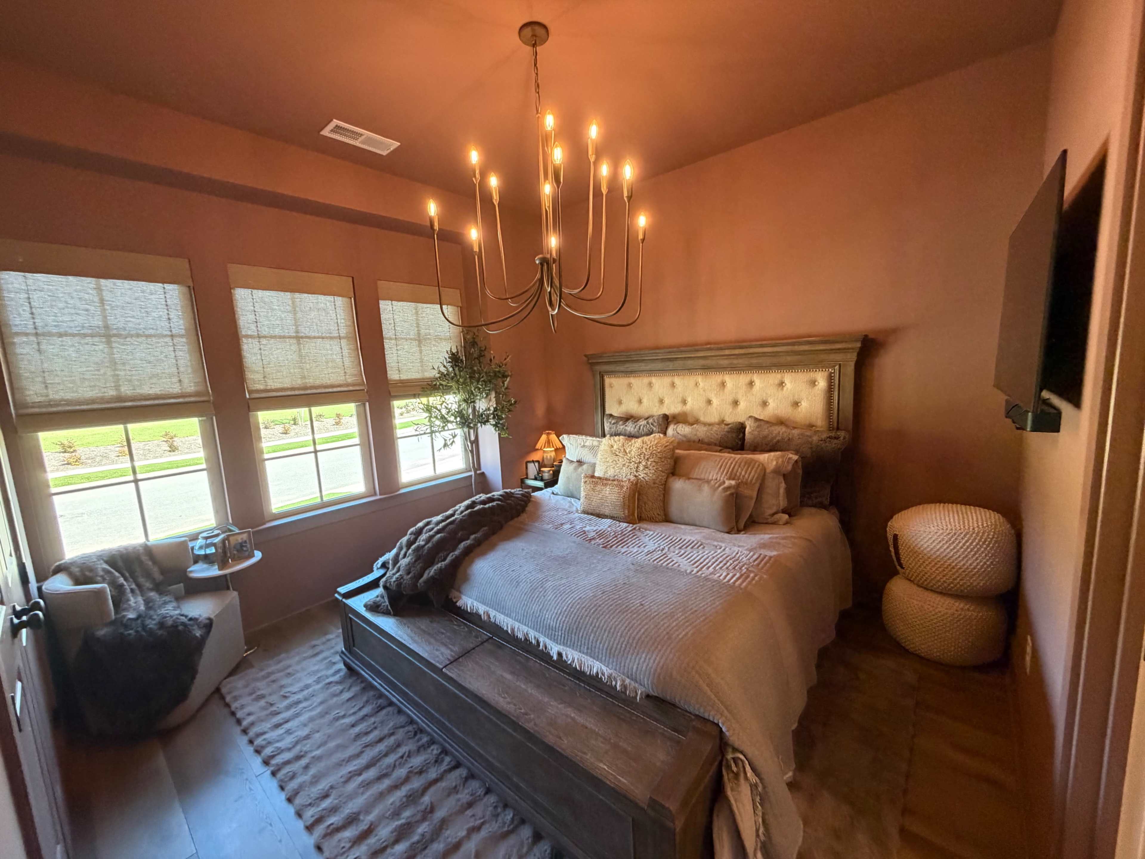 The image shows a cozy bedroom with a large bed, decorative pillows, a chandelier, and sunlight streaming through three windows.