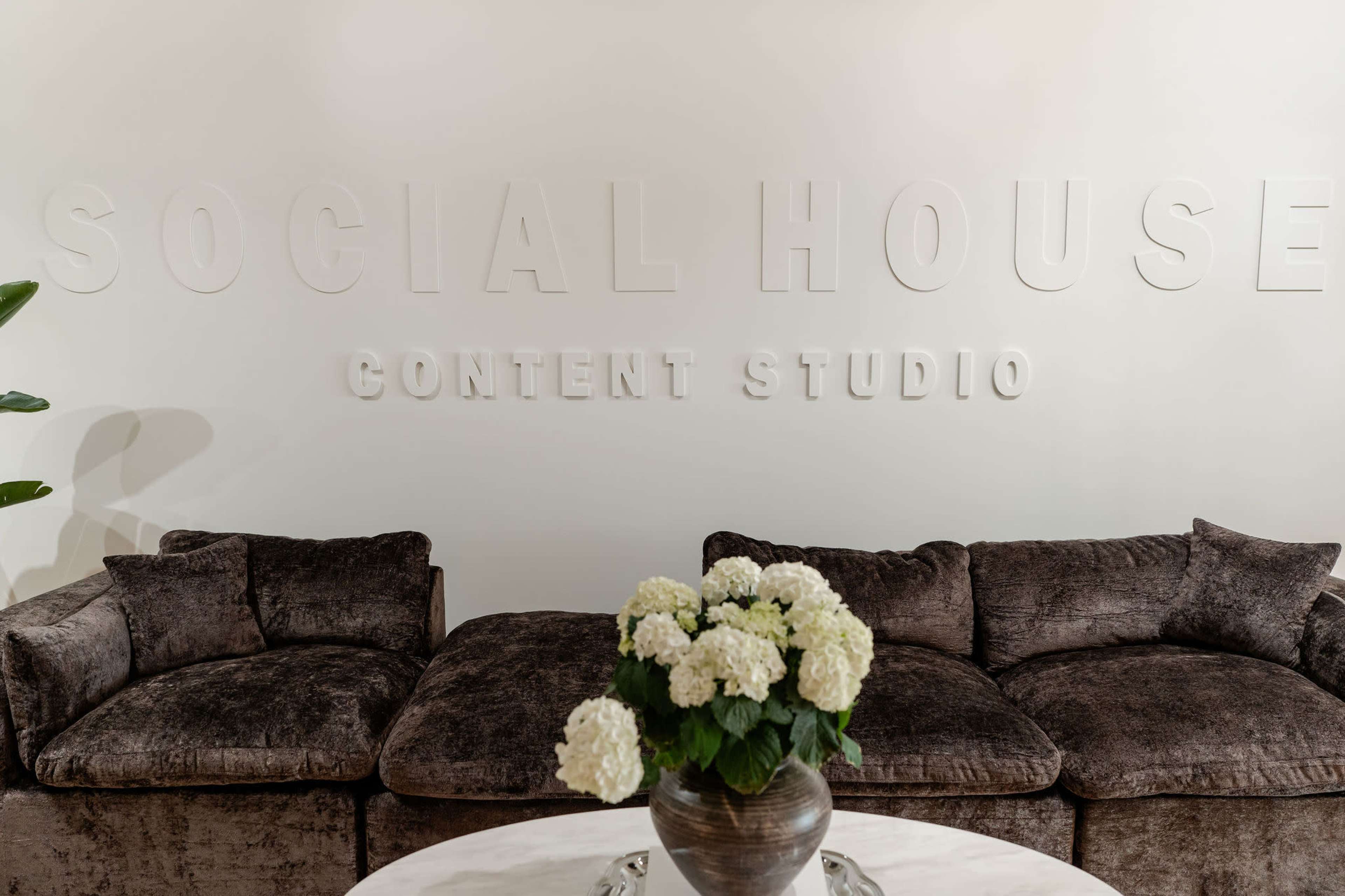 The image shows a modern interior with a dark velvet sofa set against a white wall featuring the words "SOCIAL HOUSE CONTENT STUDIO" in raised lettering, with a round table in front holding a vase of white flowers.
