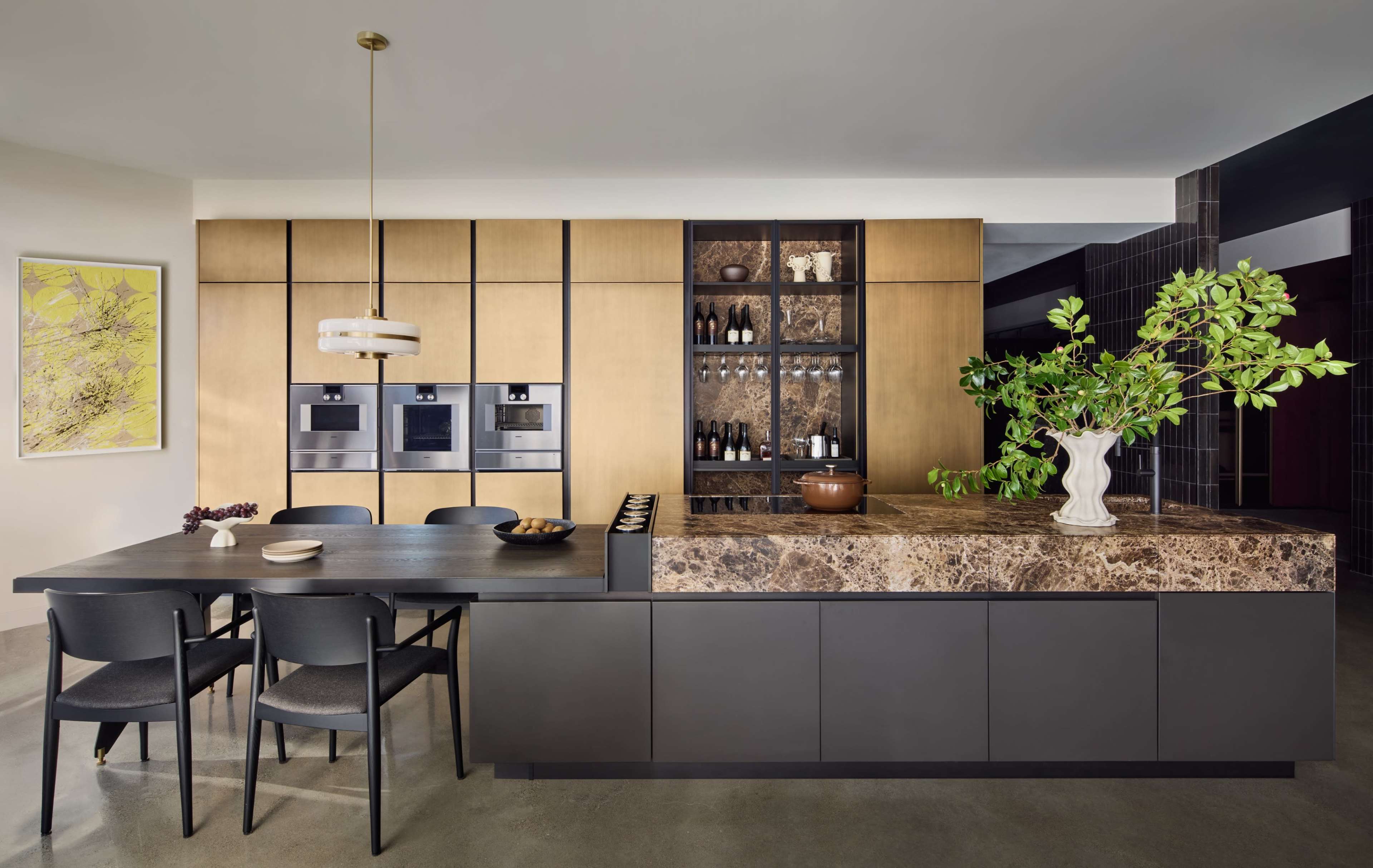 The image shows a modern kitchen with wooden cabinetry, a large dark countertop, a dining table with black chairs, and built-in appliances.