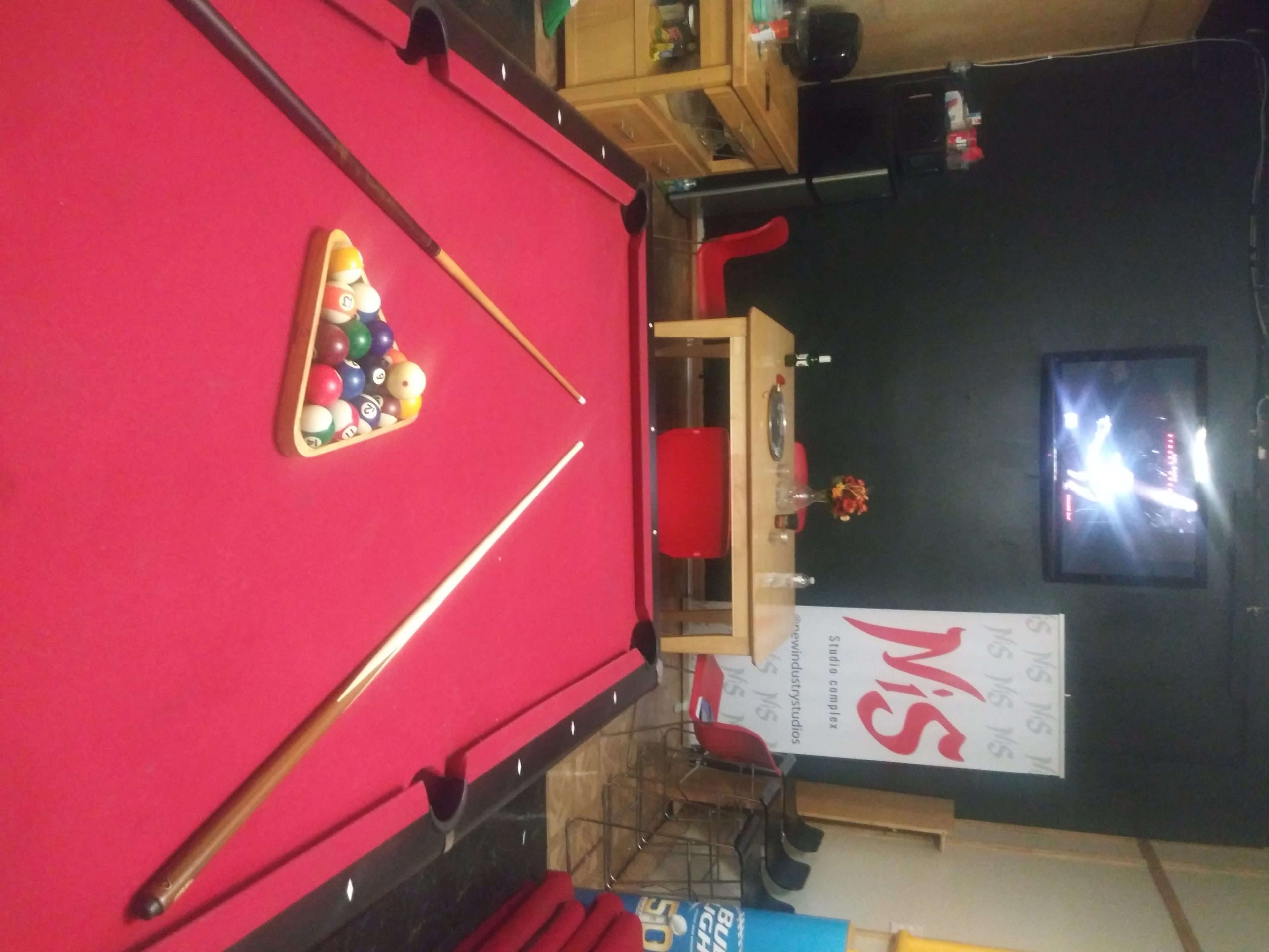 The image shows a pool table with a triangular rack of colored balls and two pool cues, with a television and dining area in the background.