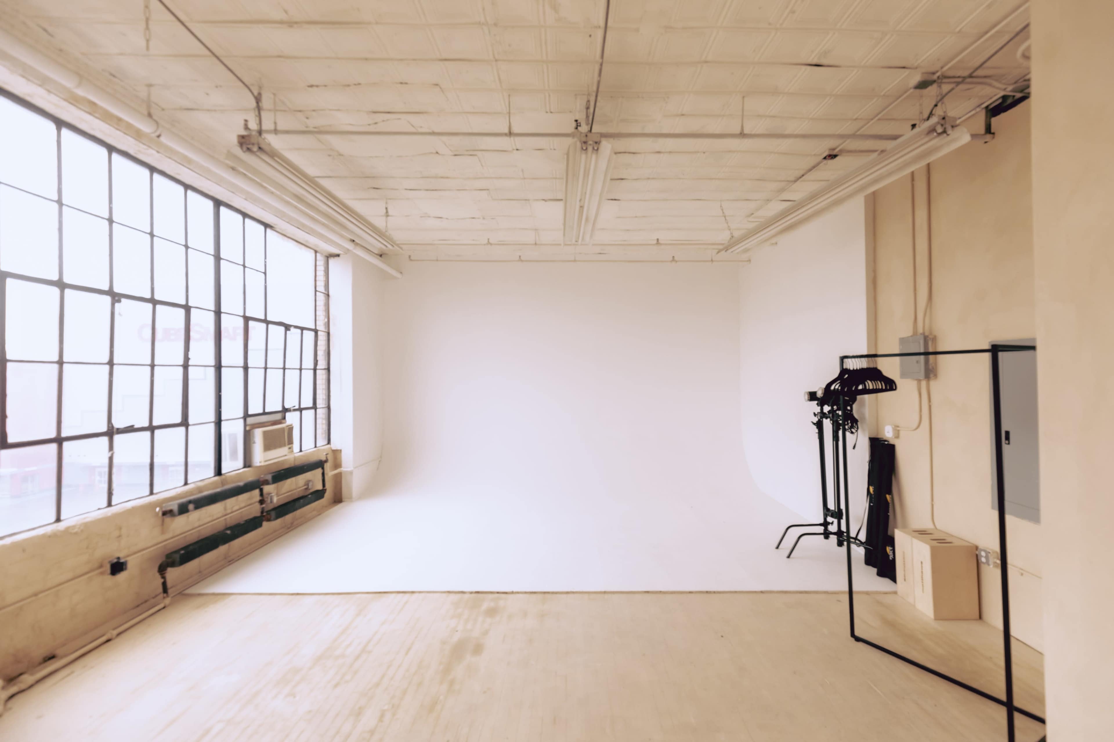 A spacious, empty studio with large windows, a plain white wall, and a clothing rack next to a mirror.