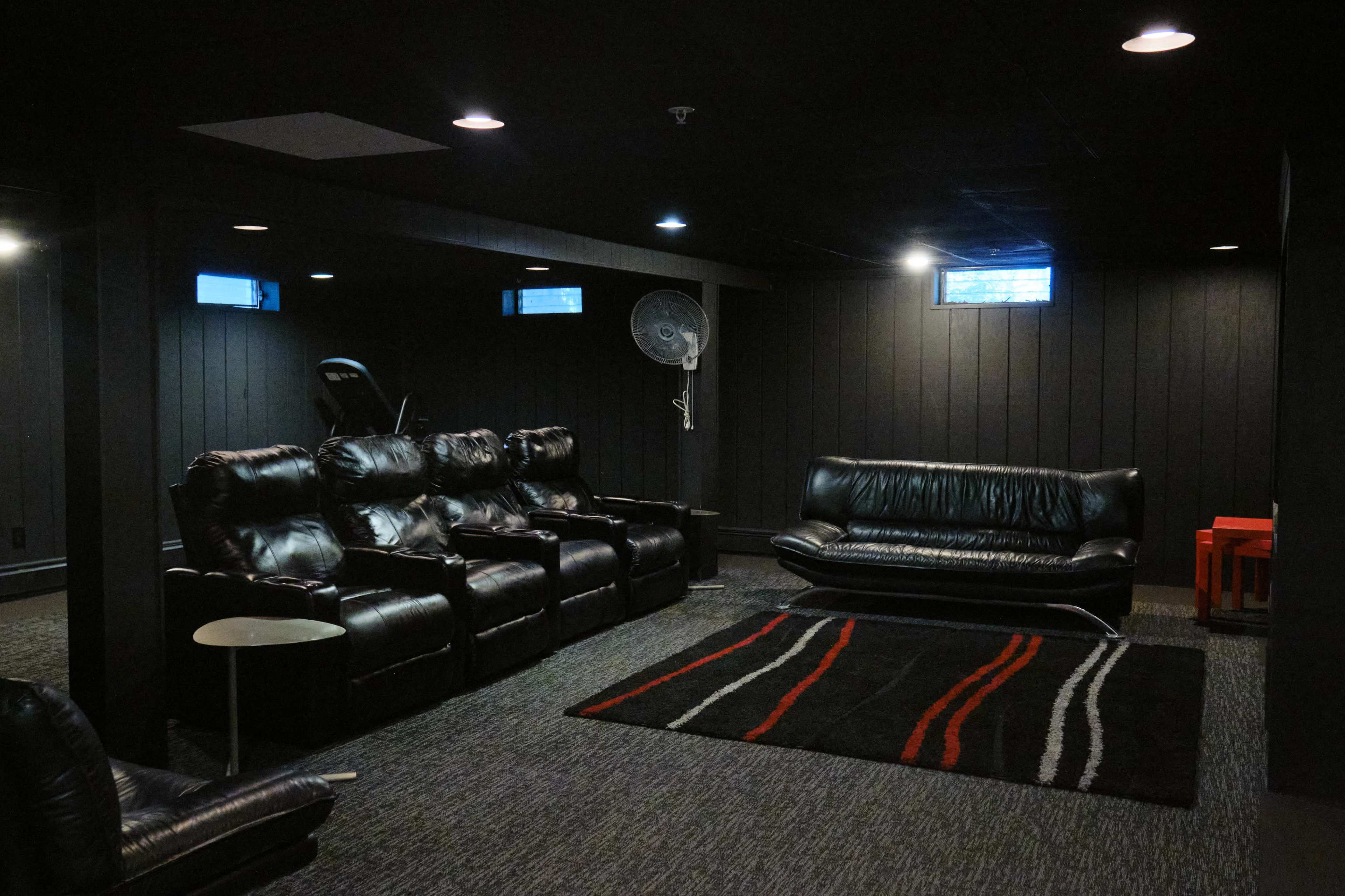 A darkly painted basement features black leather seating, a red accent table, and minimal lighting with two small windows.