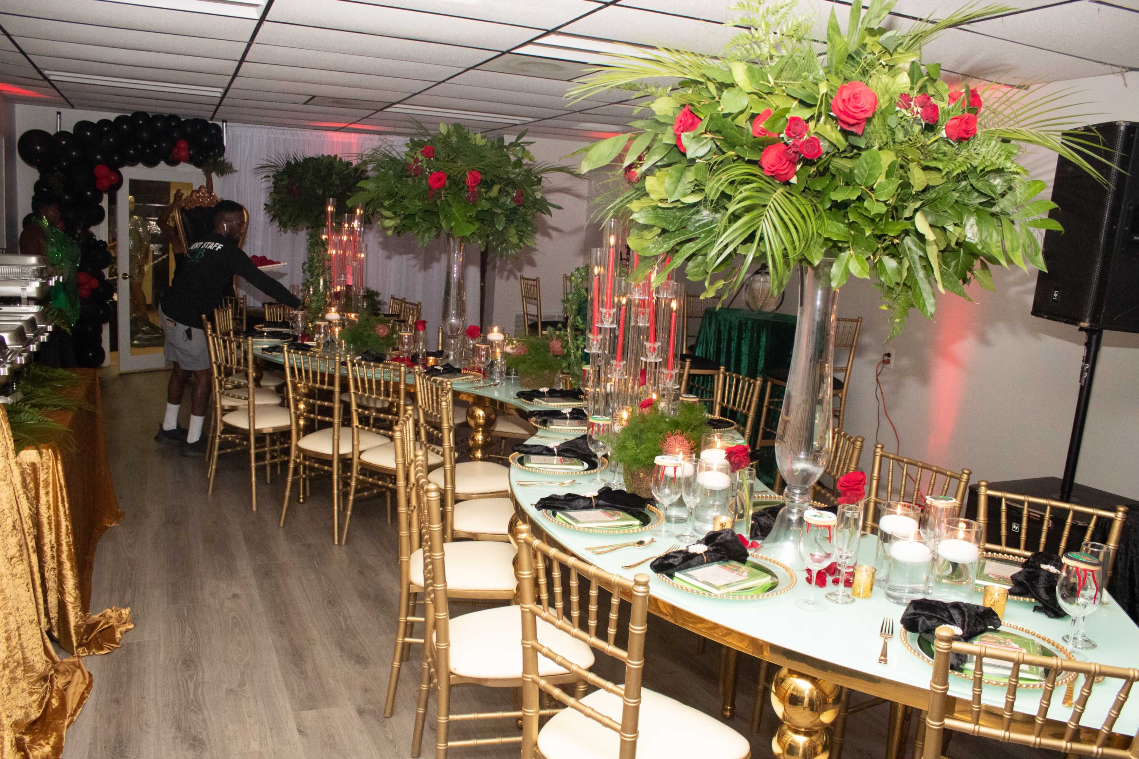 A long, elegantly set banquet table adorned with greenery and red roses is arranged in a decorated room featuring black balloons and a DJ setup.