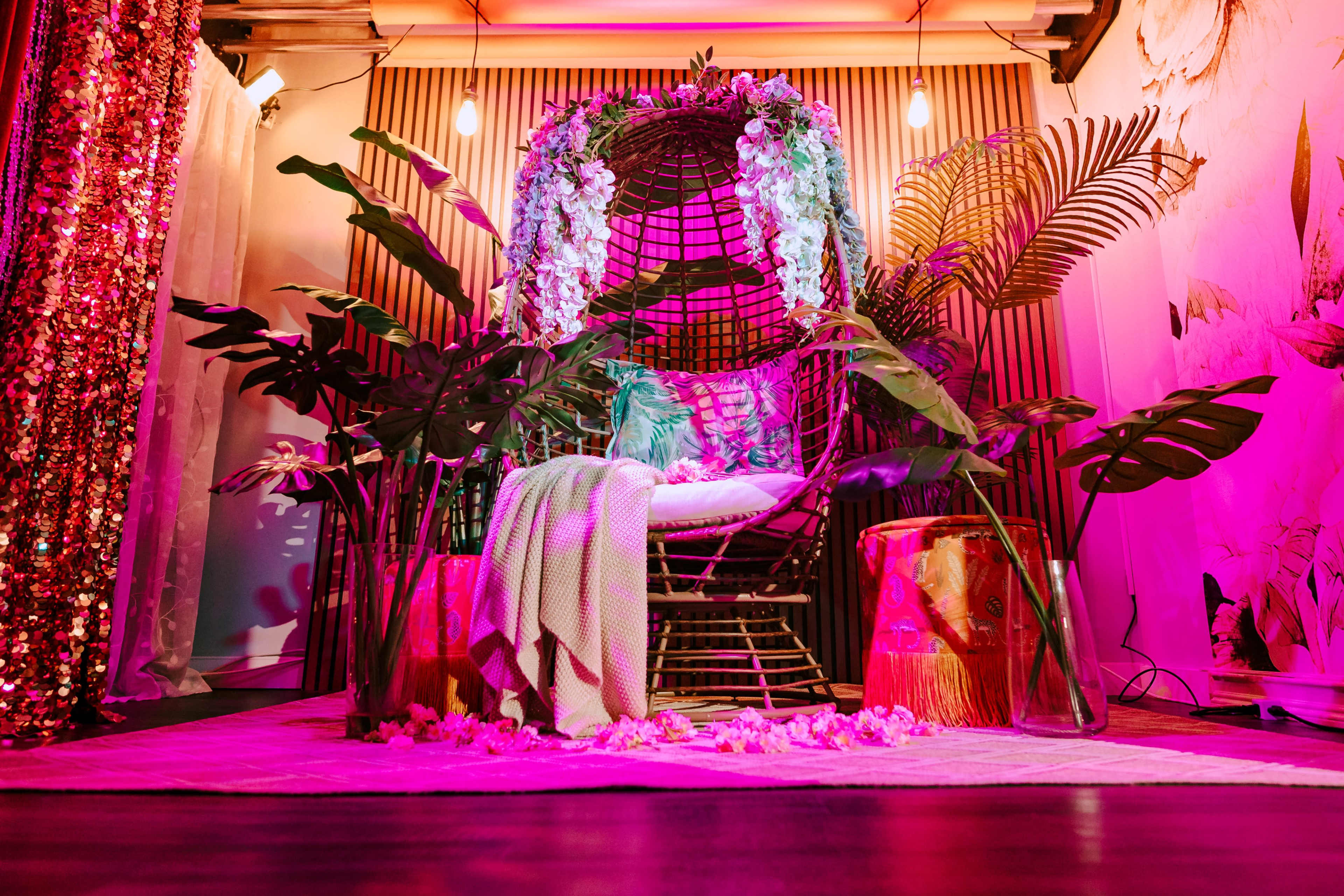 The image shows a decorative seating area featuring a circular wicker chair surrounded by vibrant foliage, colorful lighting, and elements of luxurious decor such as patterned textiles and sequined drapes.
