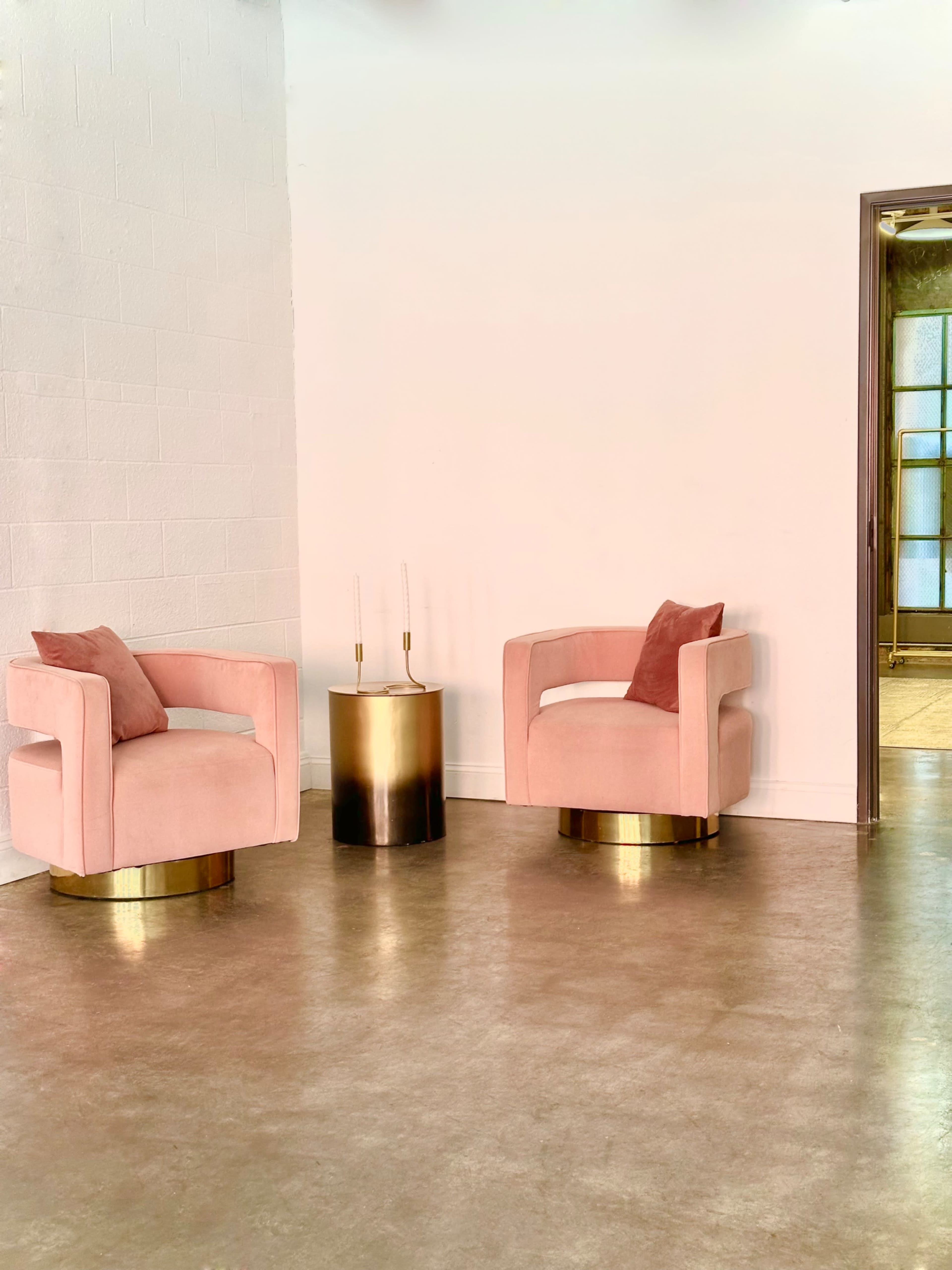 Two pink chairs with gold accents are positioned next to a round metallic side table in a minimalist room.