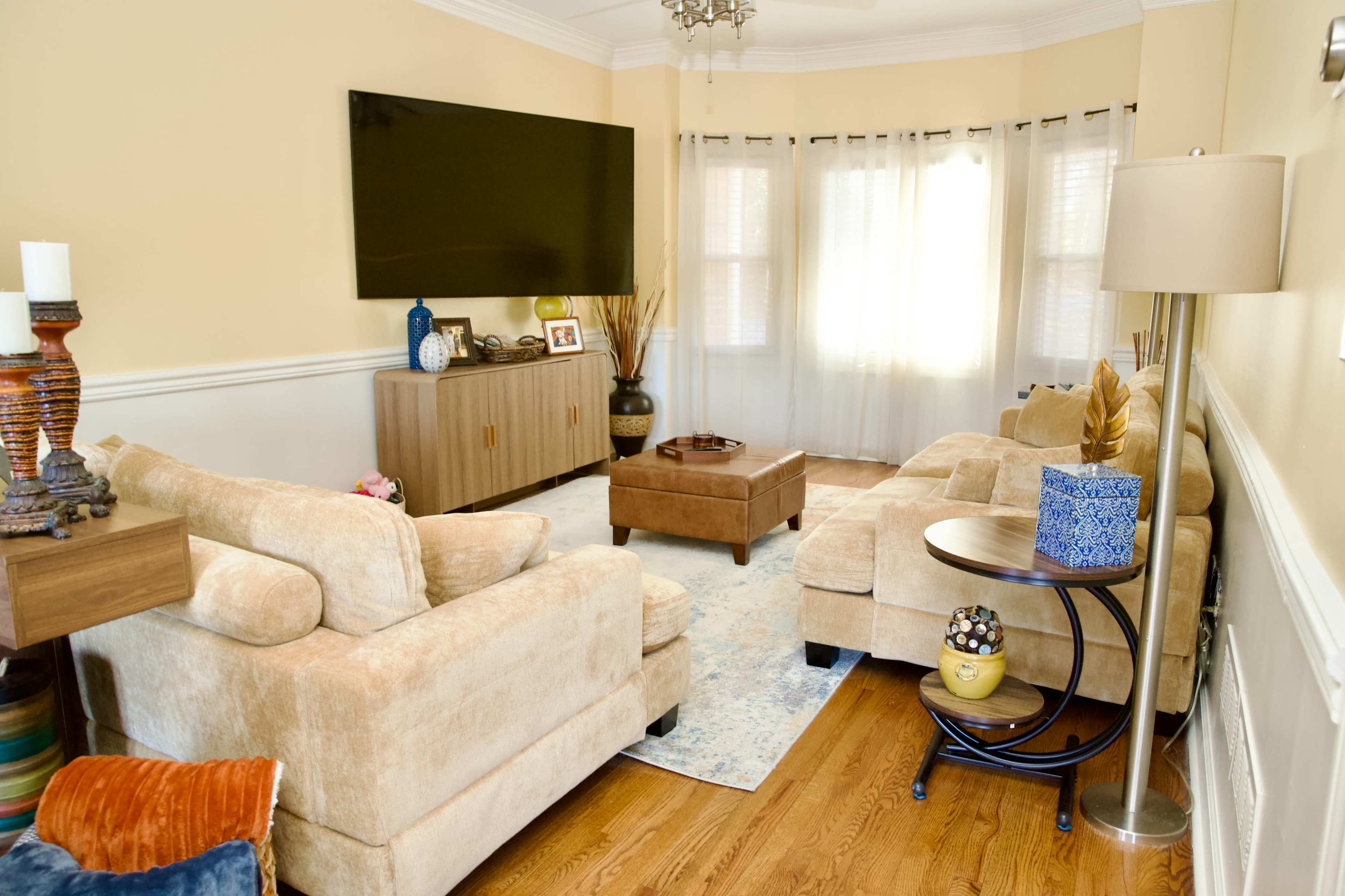 The image shows a cozy living room with two beige sofas, a wooden coffee table, a television mounted on the wall, and large windows adorned with sheer curtains.
