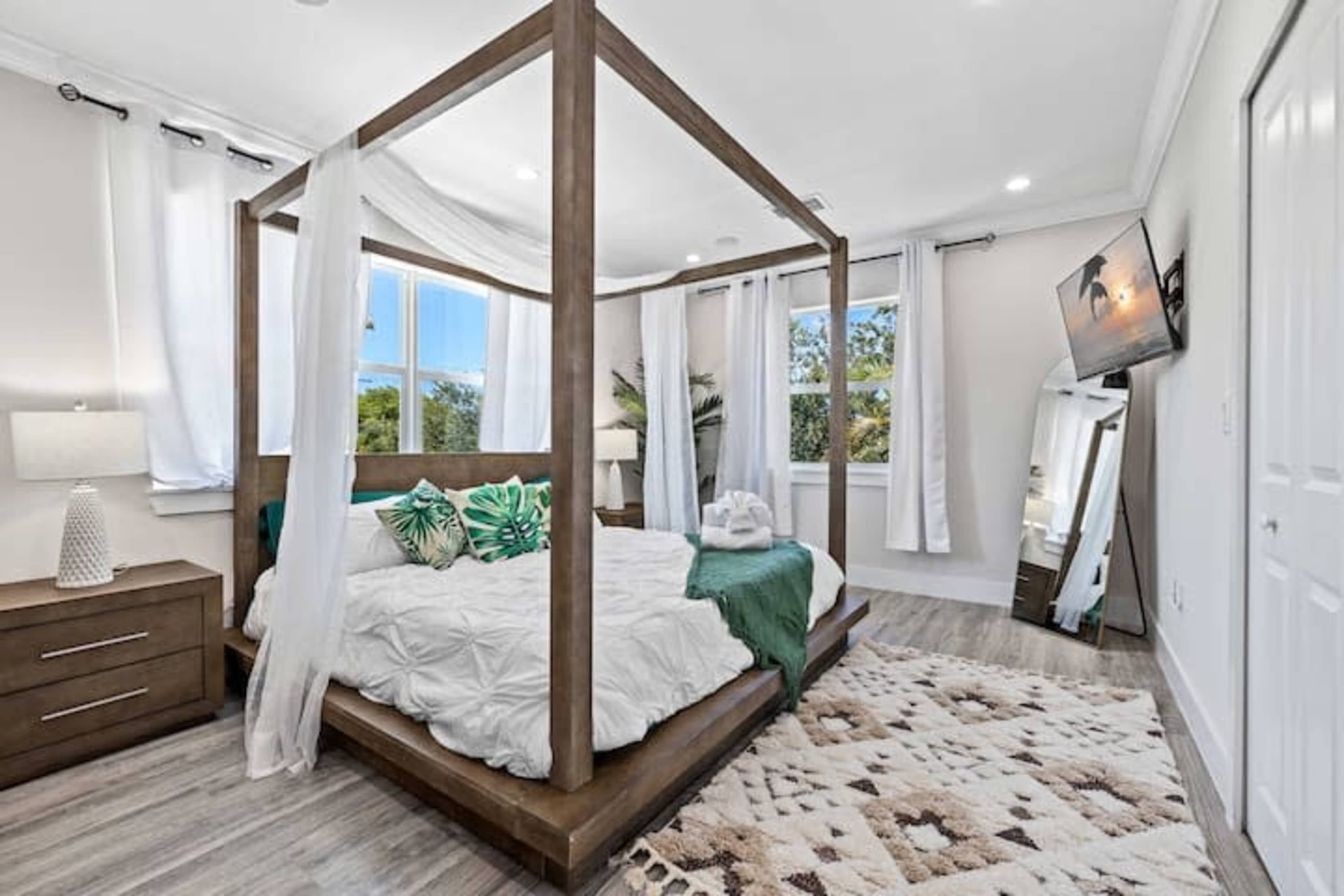 A bedroom featuring a wooden four-poster bed with light bedding, a nightstand, a large mirror, and windows with sheer curtains.