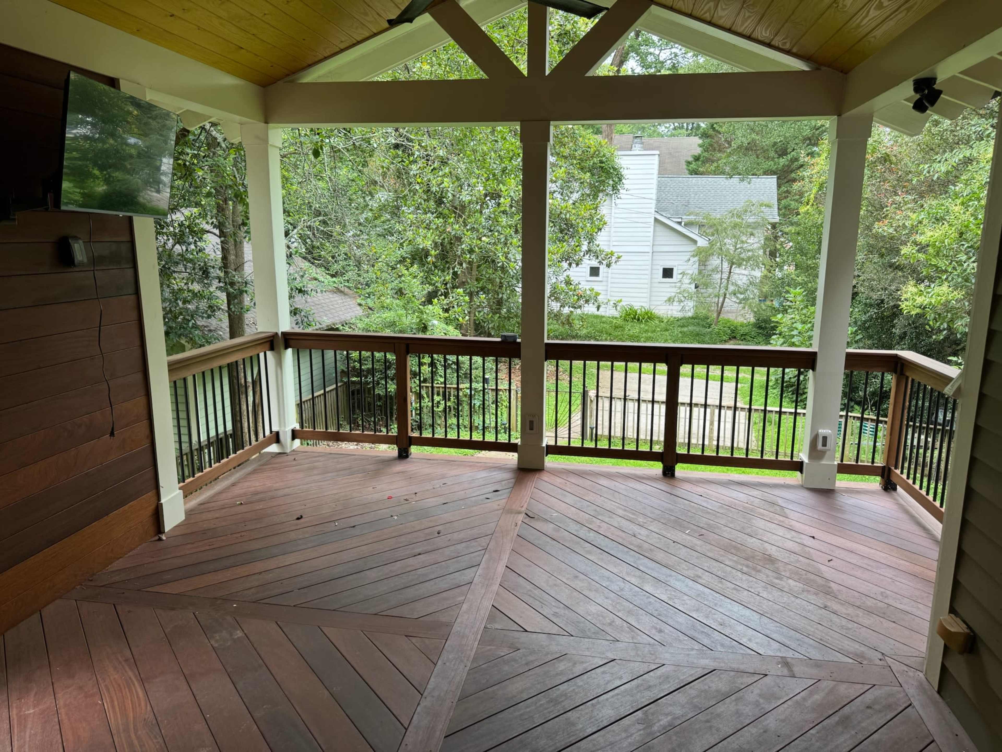 The image shows a covered outdoor deck with a wooden floor and railing, surrounded by trees and a view of nearby houses.