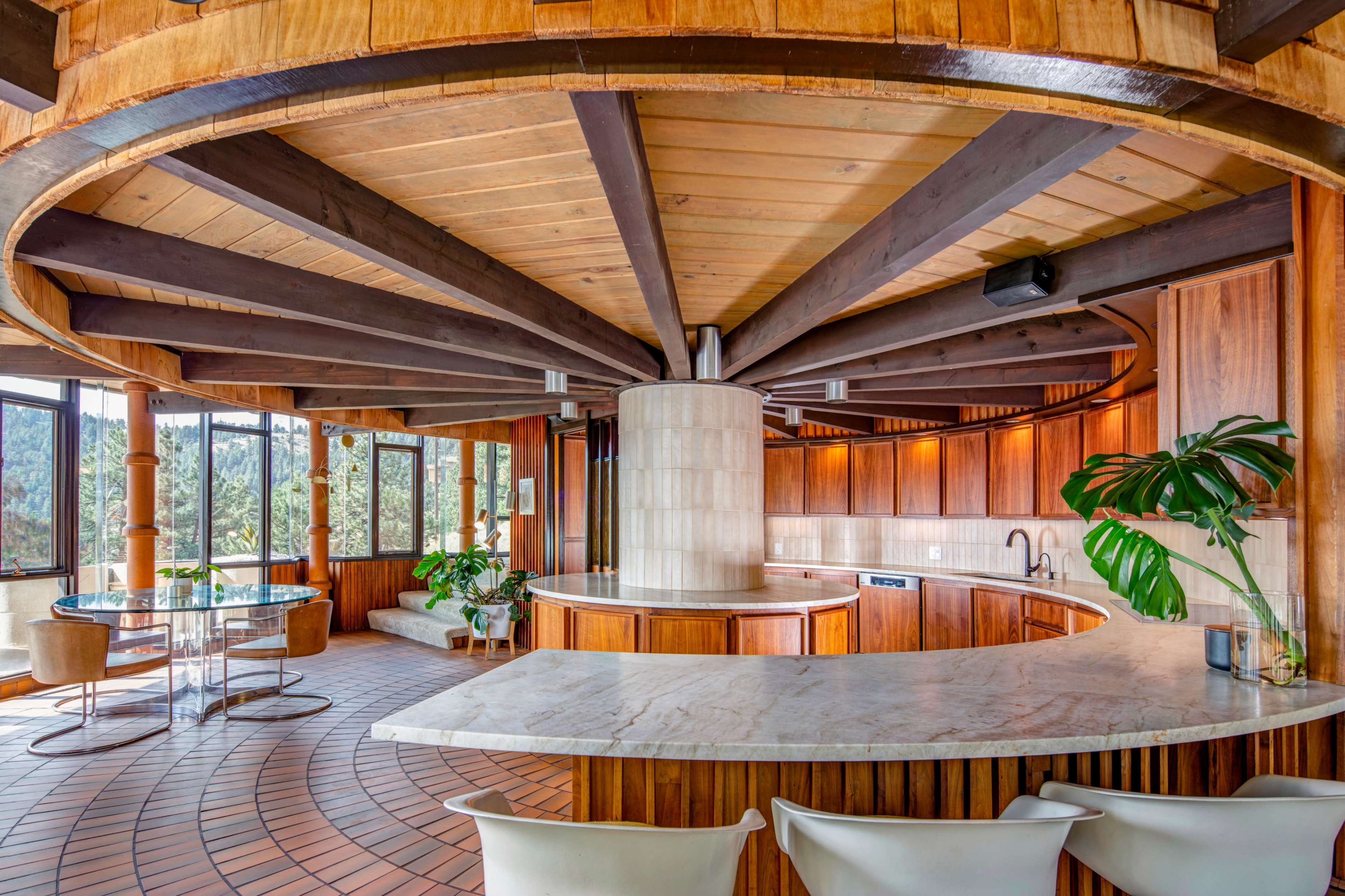 The image shows a modern, circular kitchen with wooden cabinetry, a large marble countertop, and large windows offering a view of a forested area.