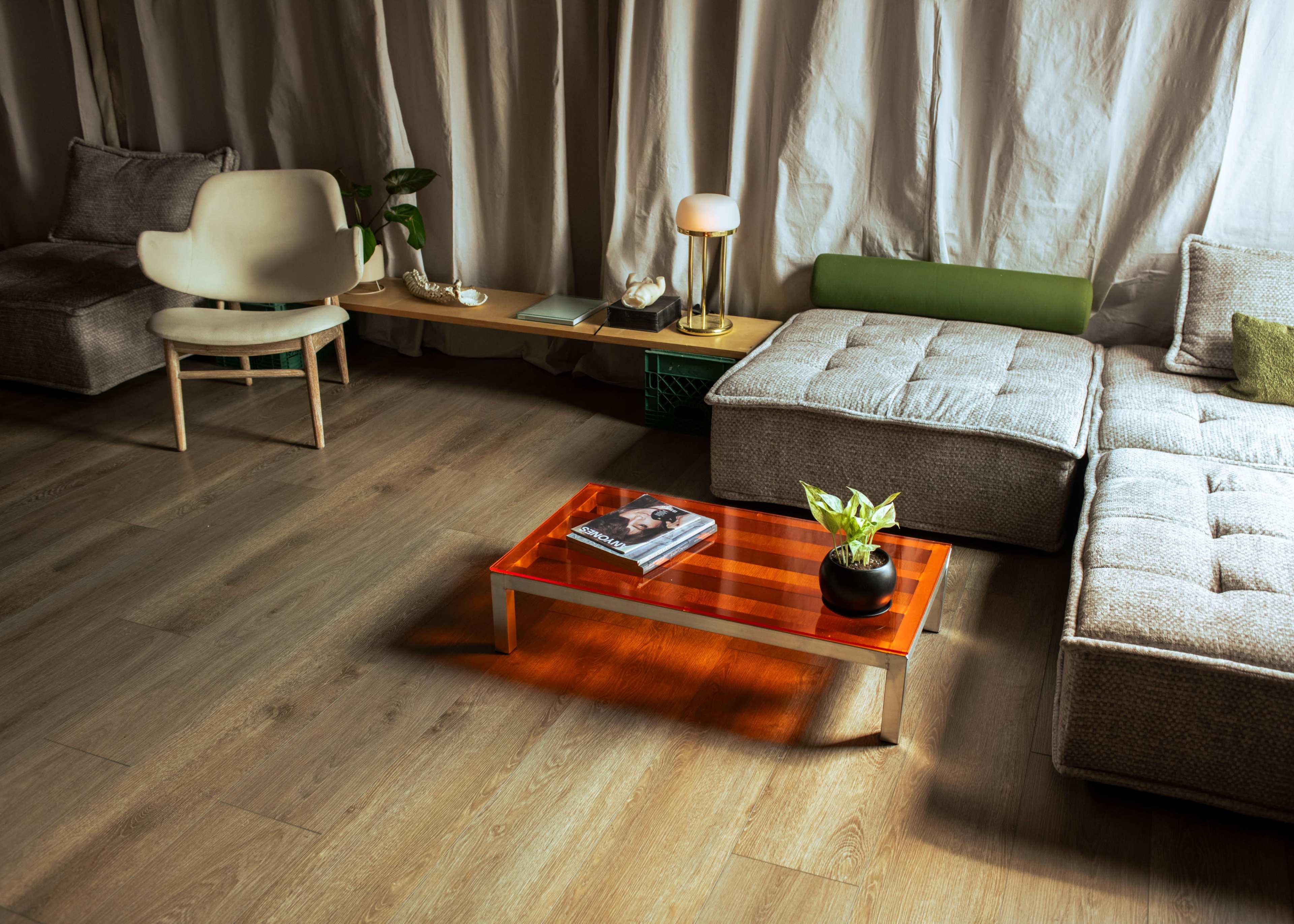 The scene features a modern living room with a textured sofa, an orange coffee table topped with a plant and a book, and a minimalist chair.