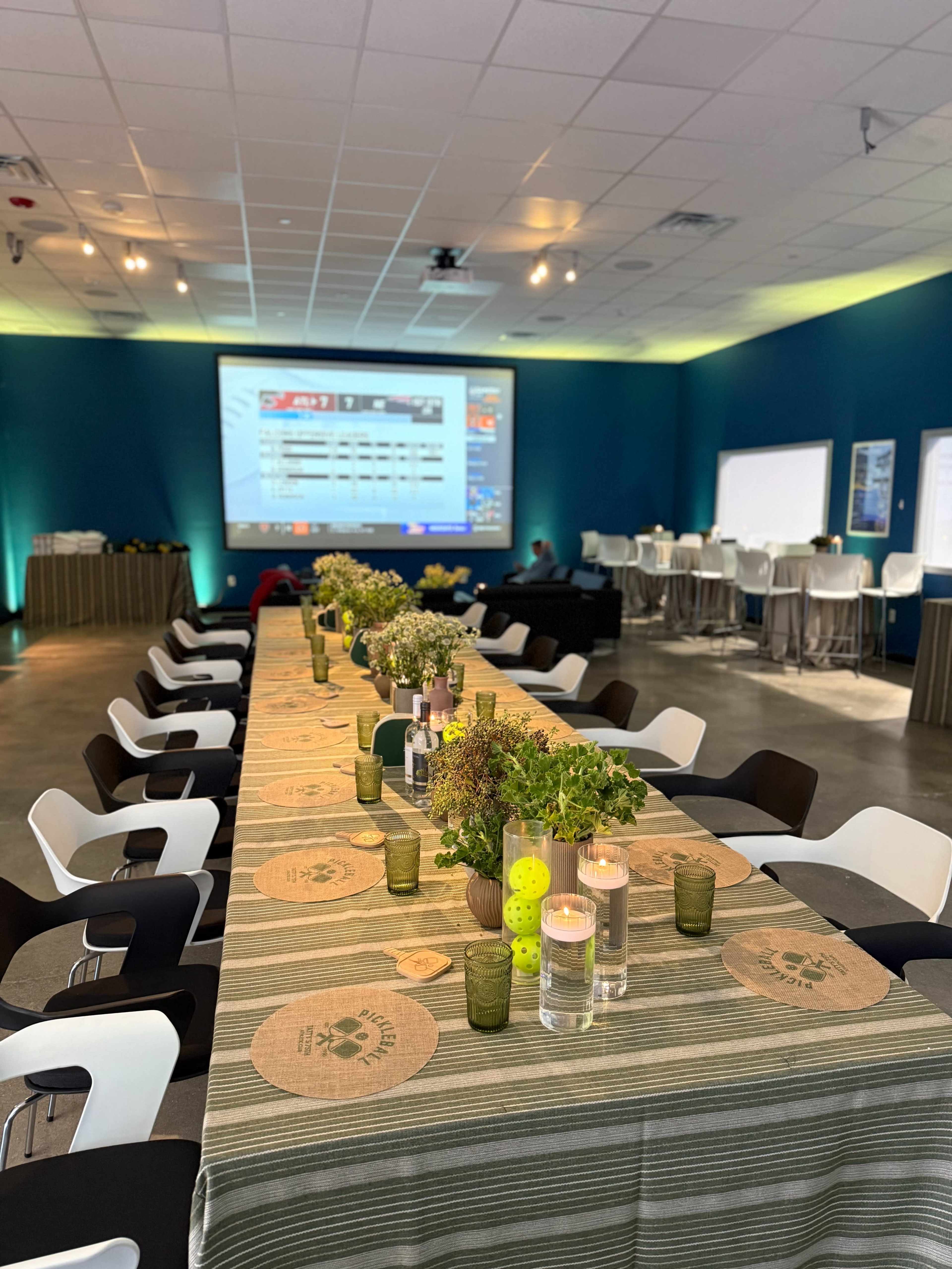 A long dining table is set with greenery and candles, with a large screen displaying information in the background and additional seating areas around the room.