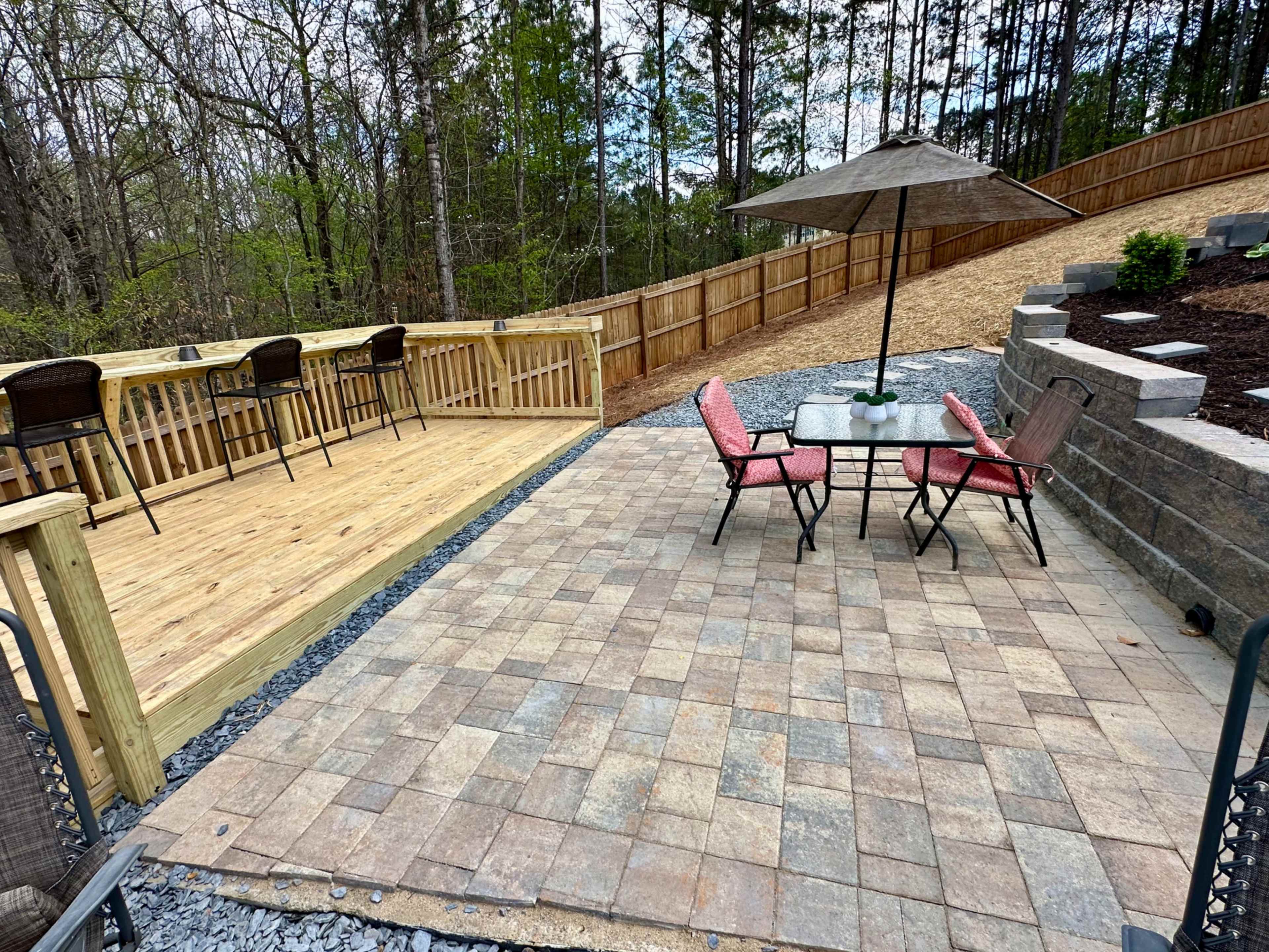 The image displays a landscaped outdoor space featuring a paver patio with a table and chairs, a wood deck with bar stools, and a sloped area lined with rocks and plants.