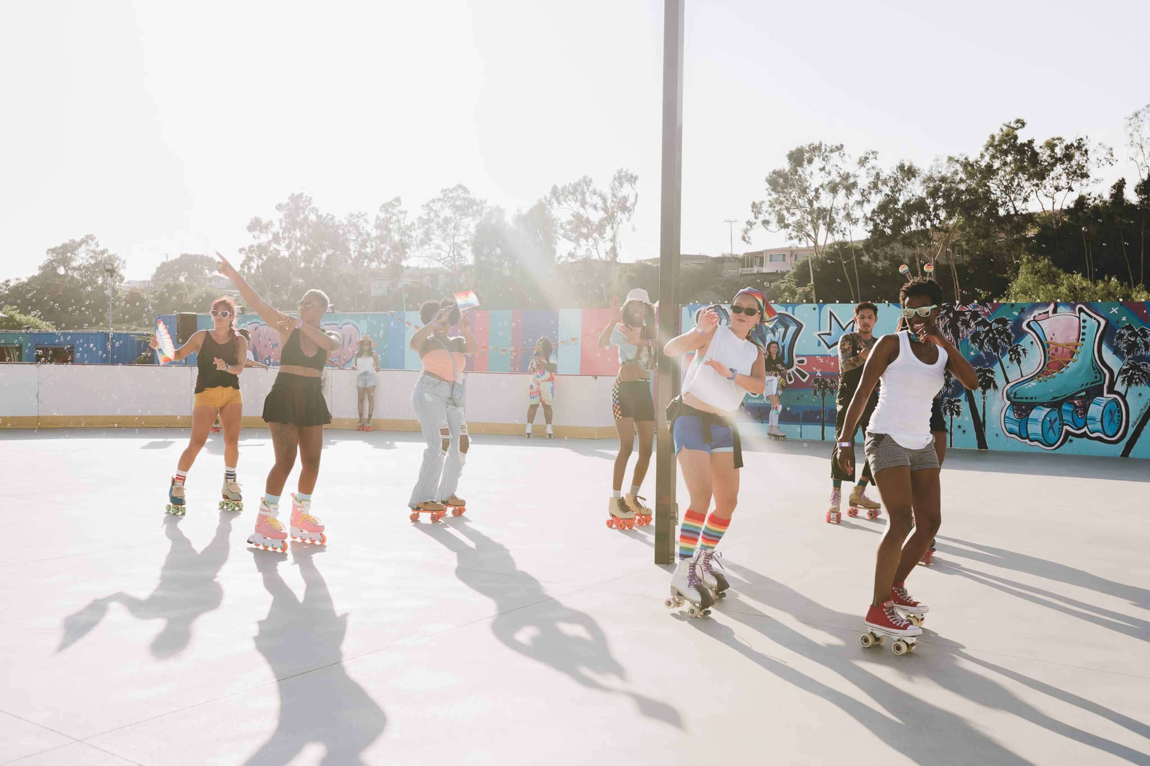 A group of people of various ages roller skate on a brightly lit rink while colorful murals decorate the surrounding walls.