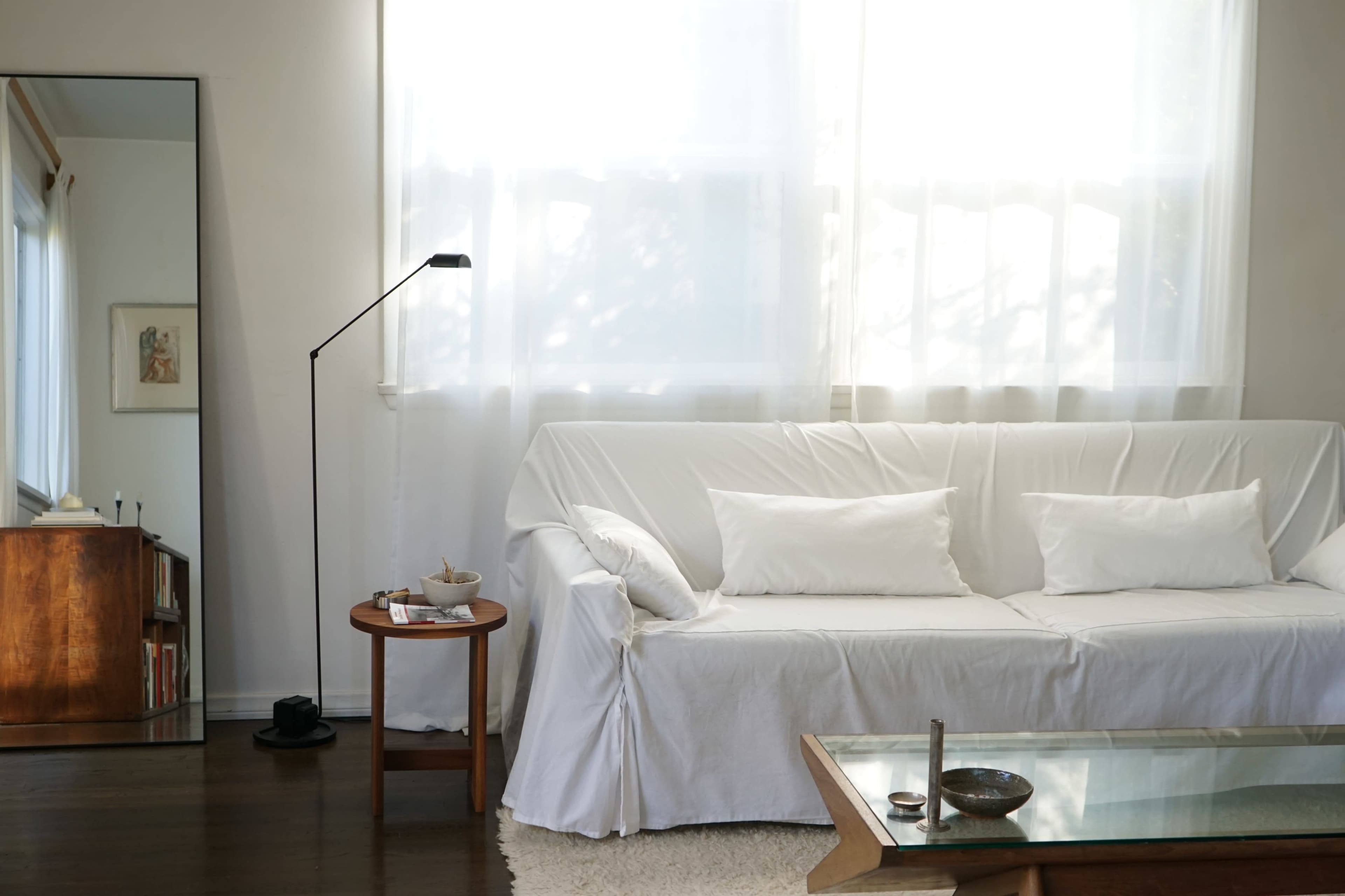 A white-covered sofa and a round coffee table are positioned in a bright living room with large windows letting in natural light.