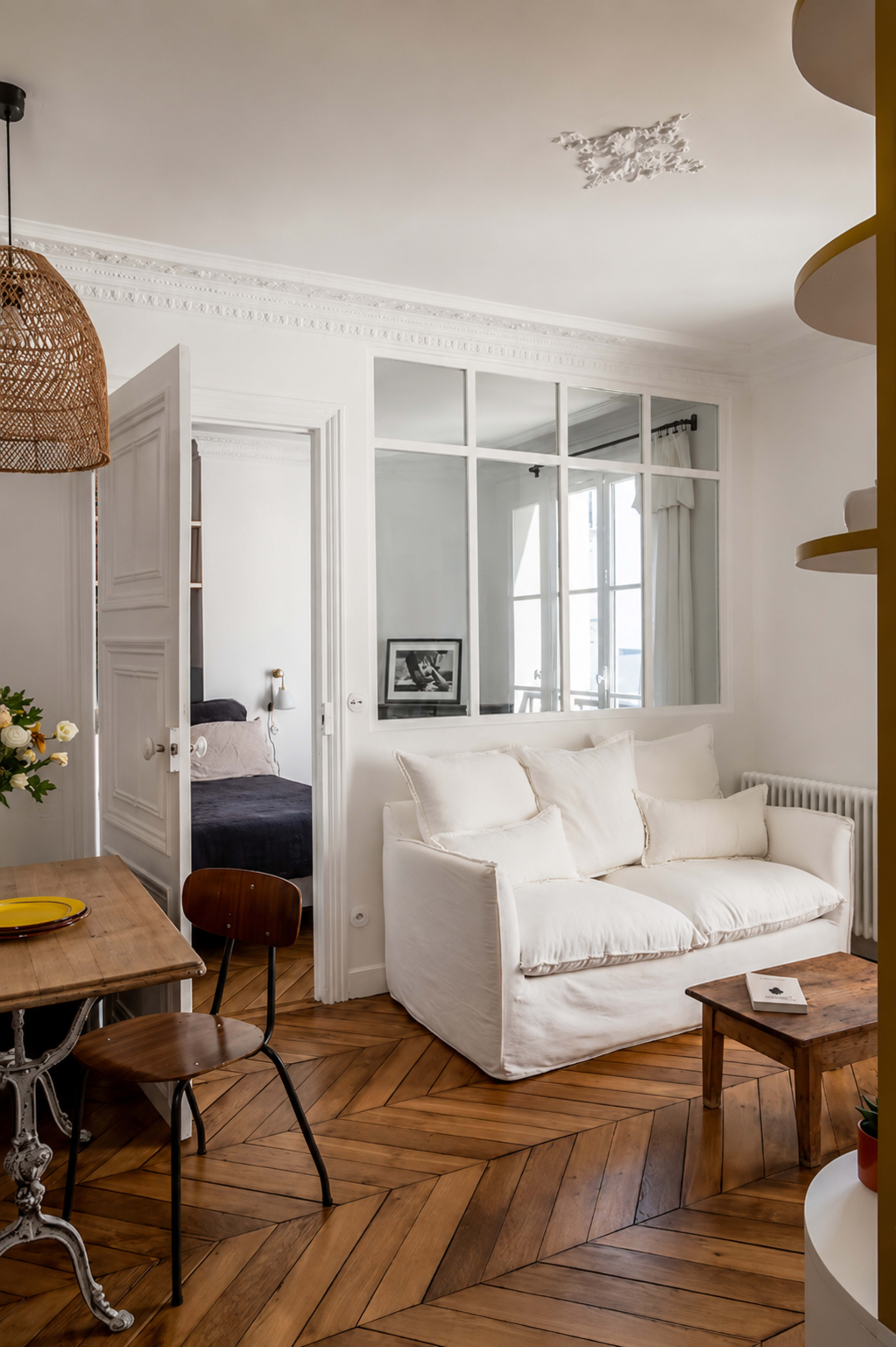 The image features a cozy living space with a white sofa, a wooden dining table, and large glass-paneled doors separating it from a bedroom.