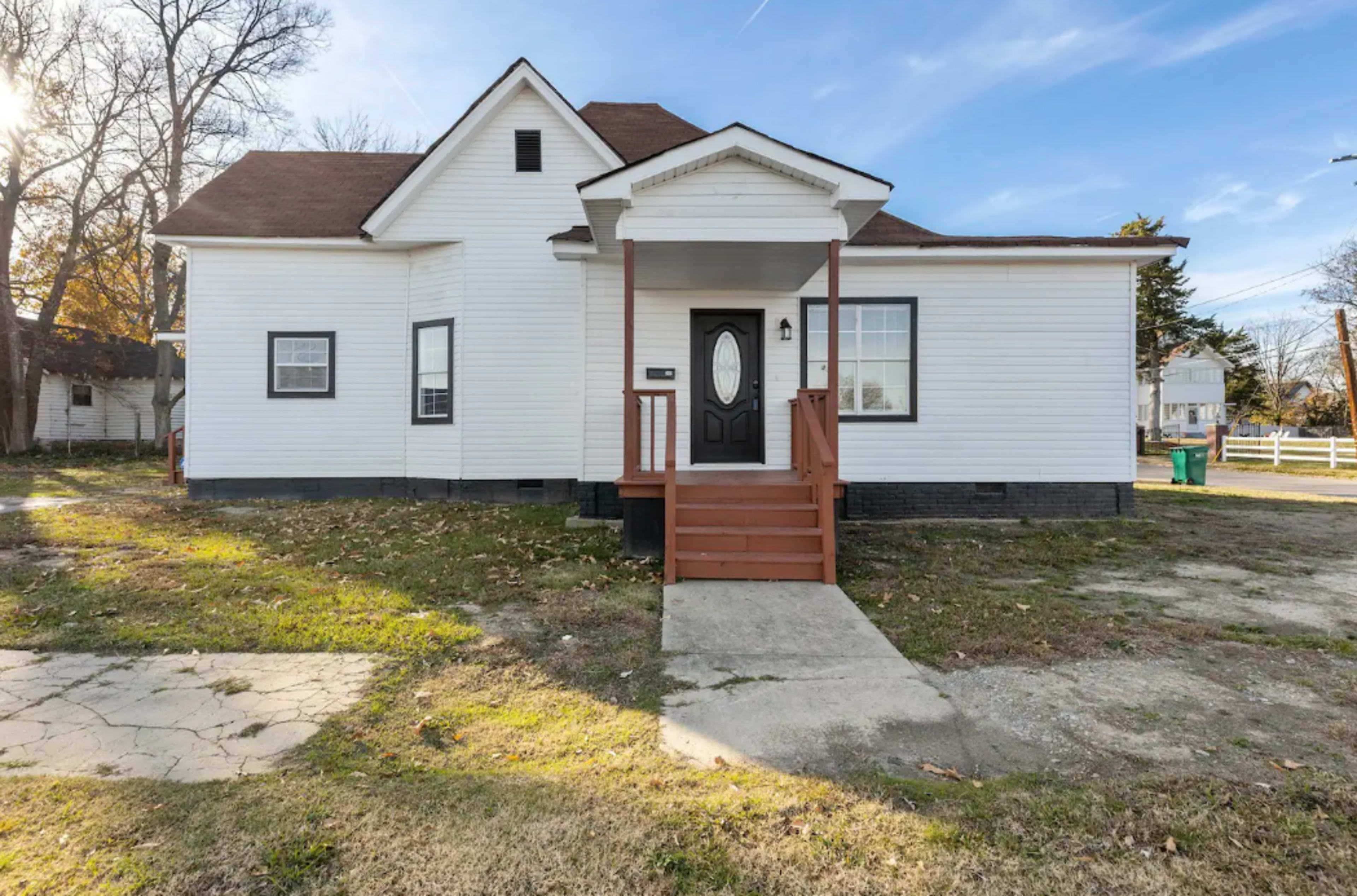 A white, two-story house with a brown roof and a front porch, surrounded by a lawn with scattered leaves and a concrete walkway.