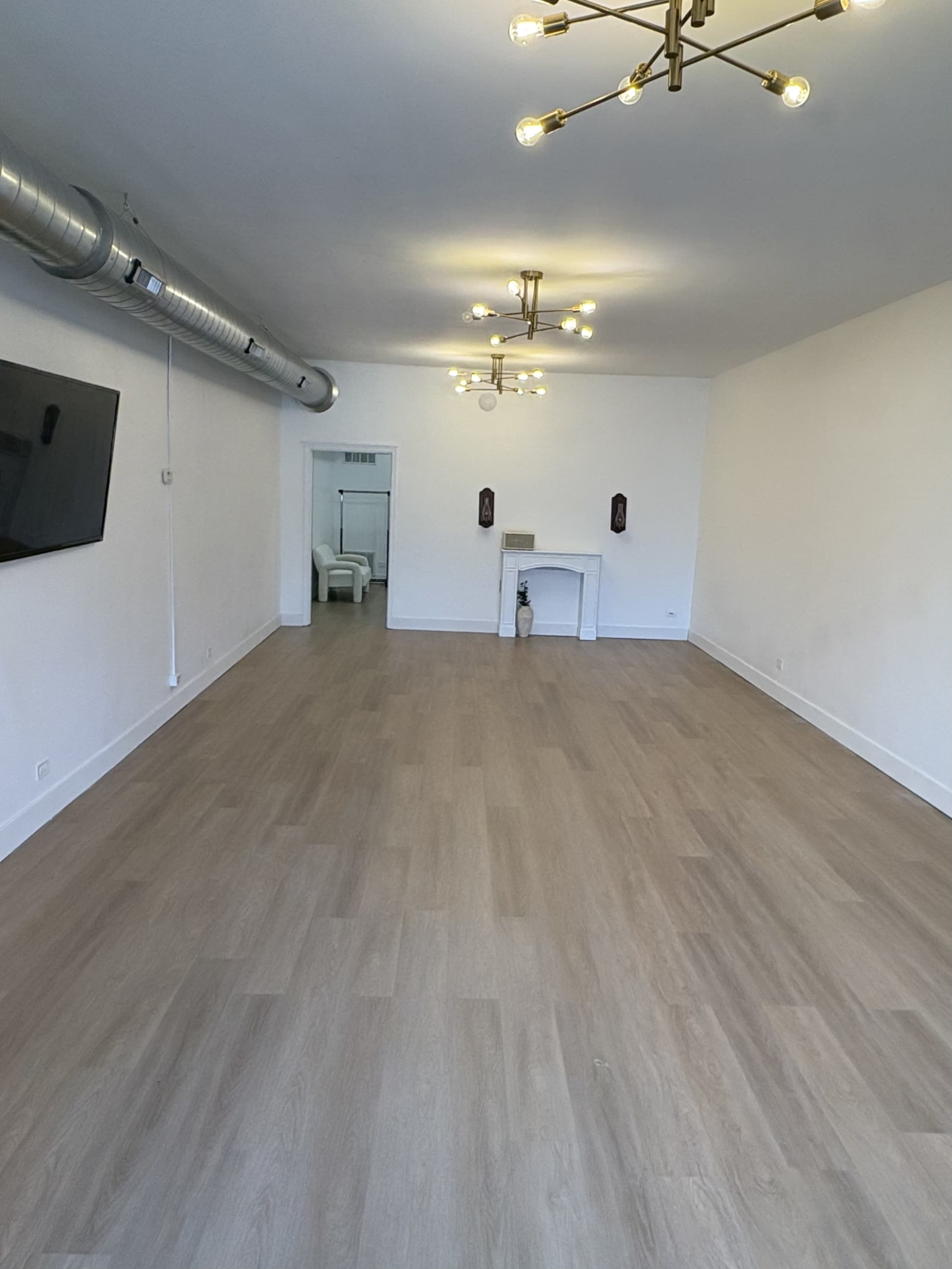 The image shows a spacious, empty room with wooden flooring, a small fireplace, and modern light fixtures on the ceiling.