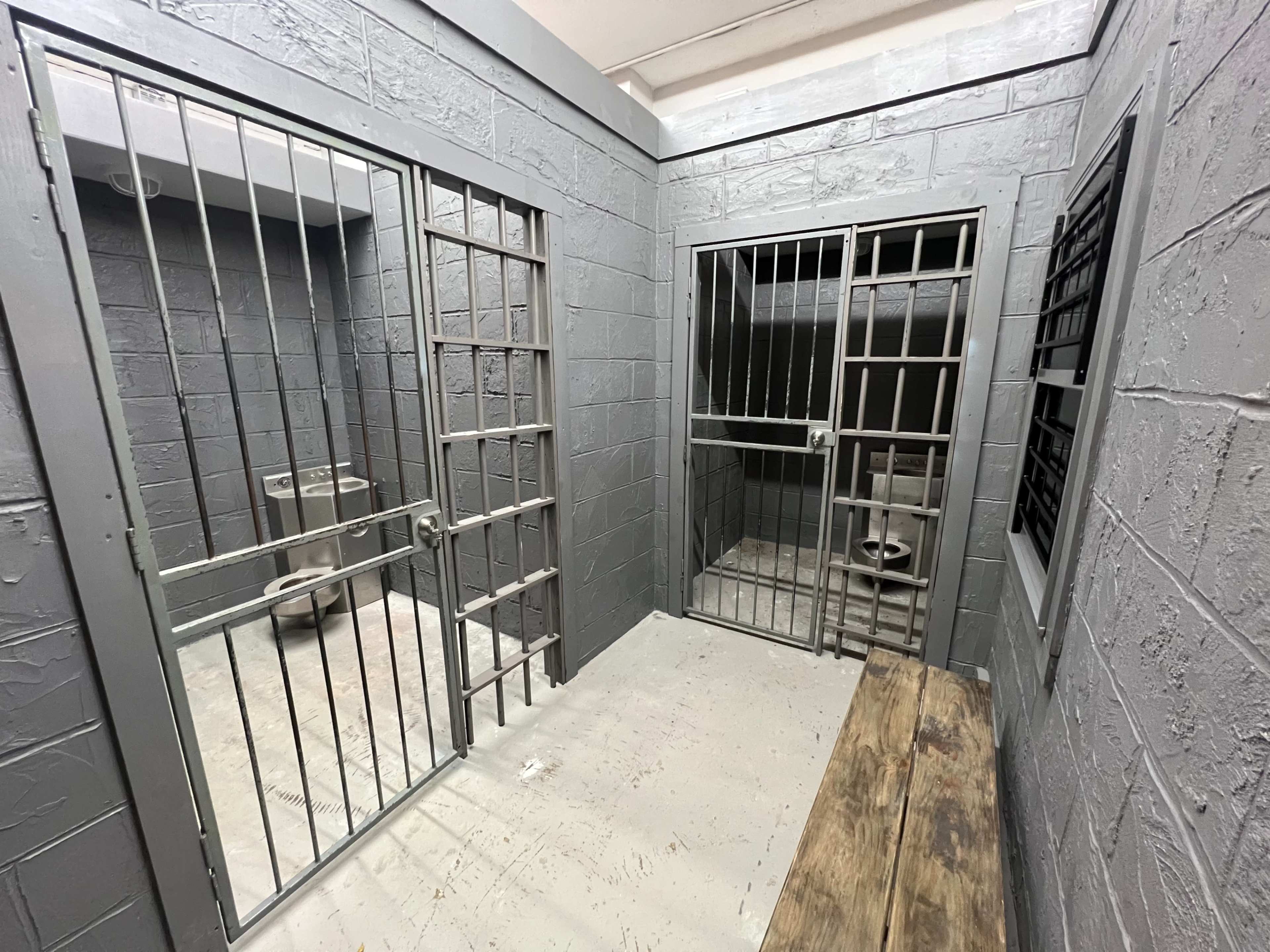The image shows a gray-walled jail cell with two barred doors, one open and one closed, and a bench along the wall.