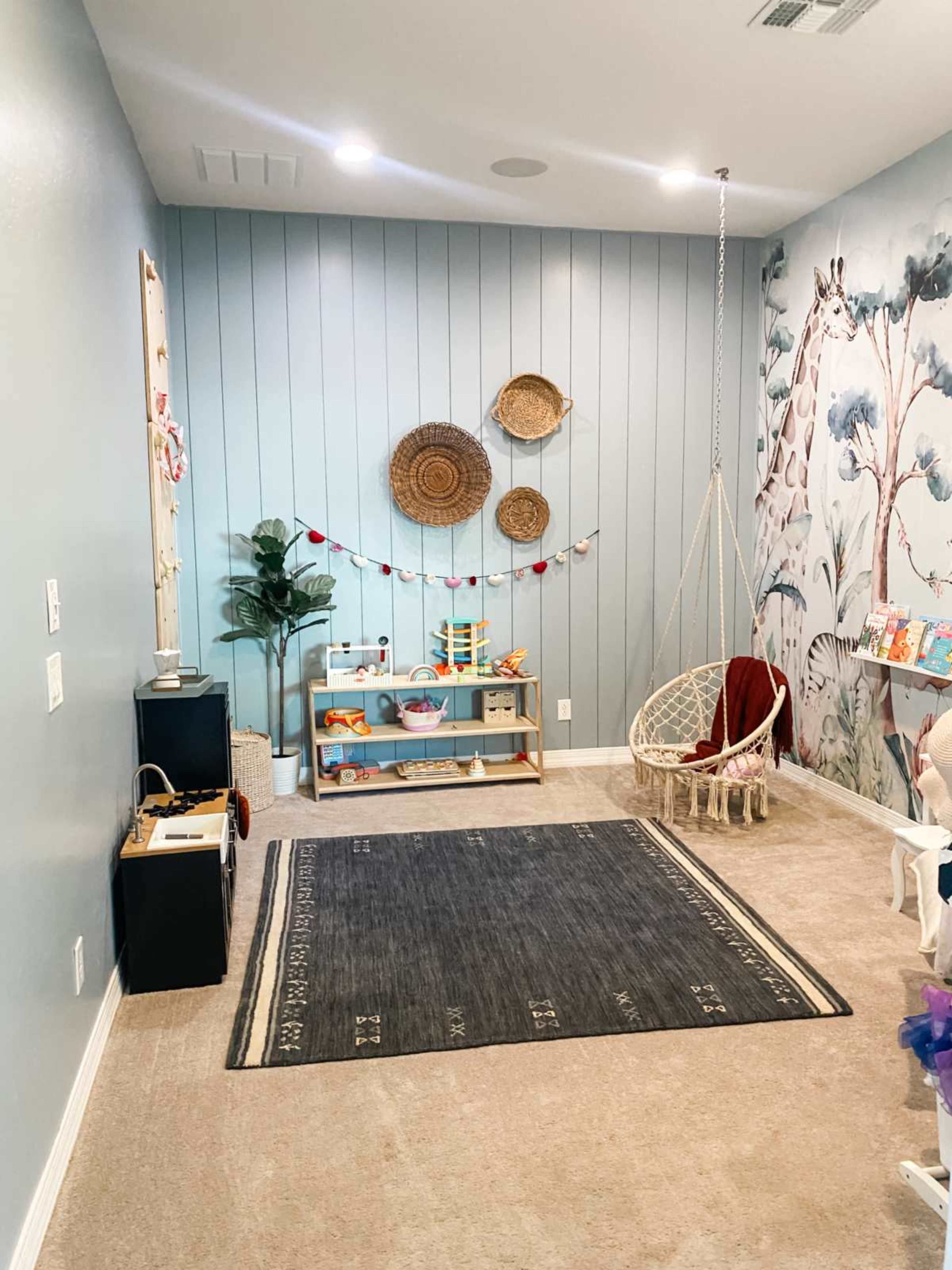 The room features light blue walls with decorative baskets on one side, a macramé hanging chair, a small bookshelf filled with books, and a neutral area rug on the carpeted floor.