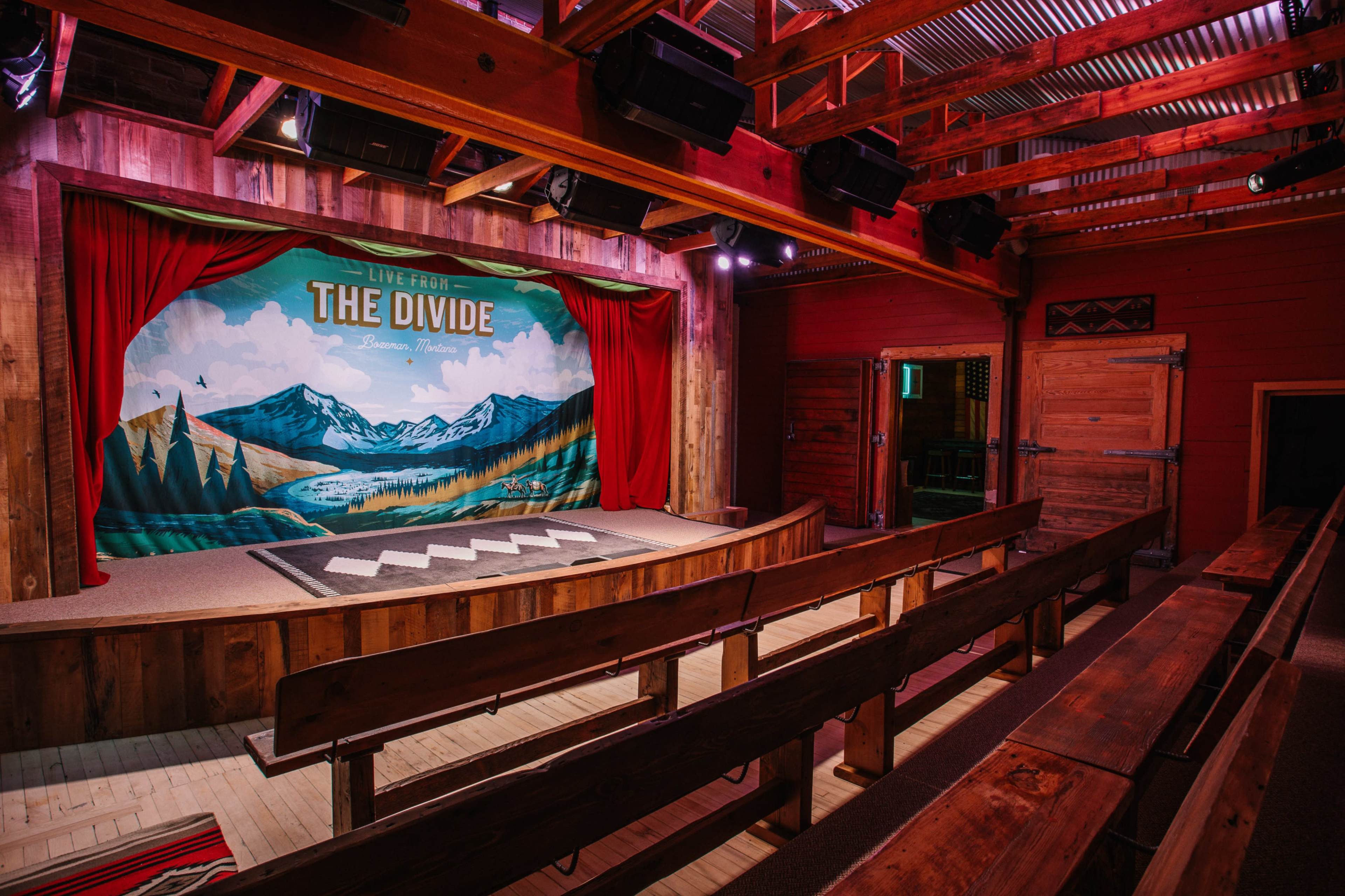 A wooden stage with a painted backdrop of mountains and trees, flanked by red curtains, set in a rustic venue with wooden seating.