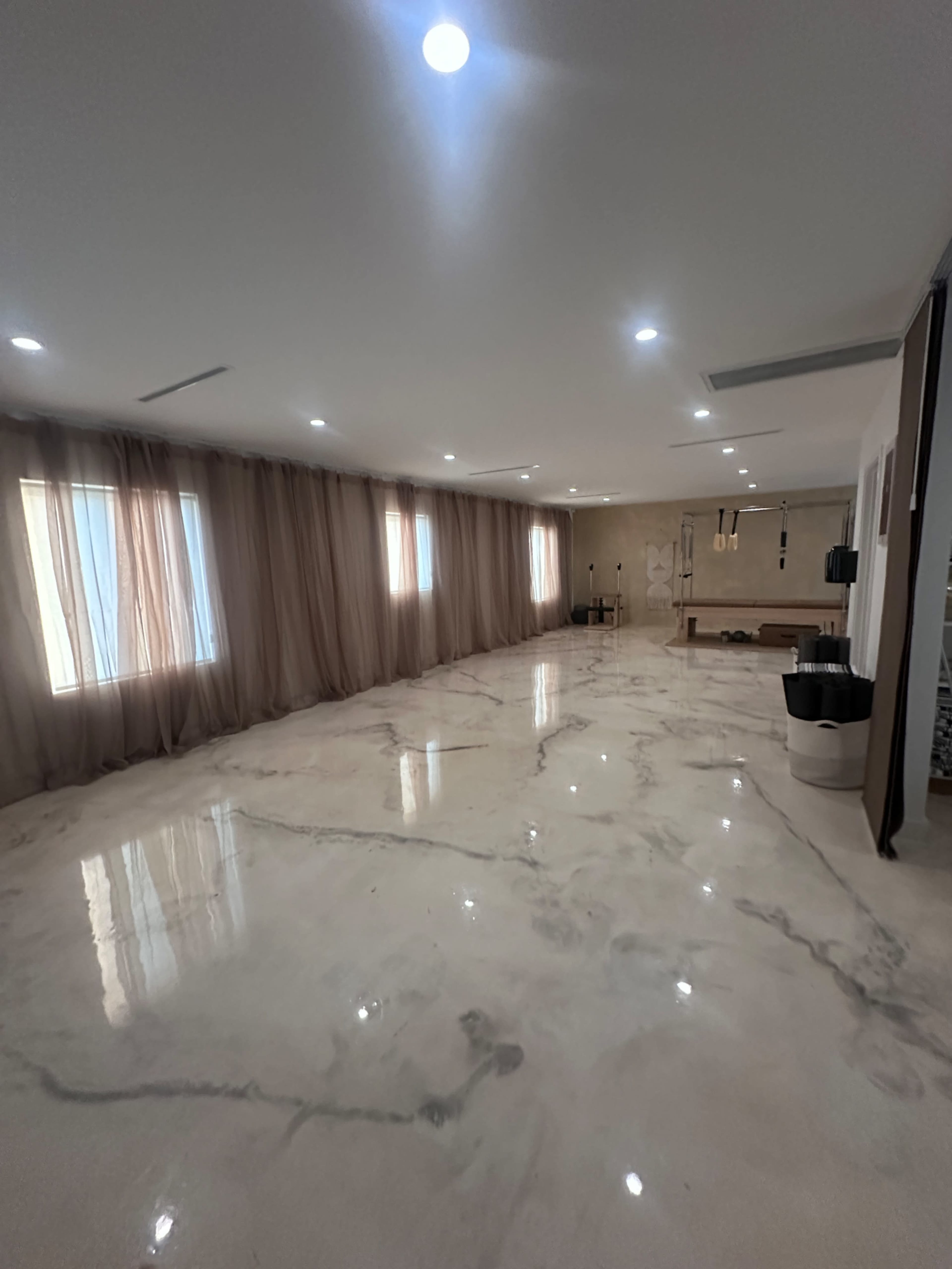 The image shows a spacious, well-lit room with marble flooring and large windows draped in light curtains.