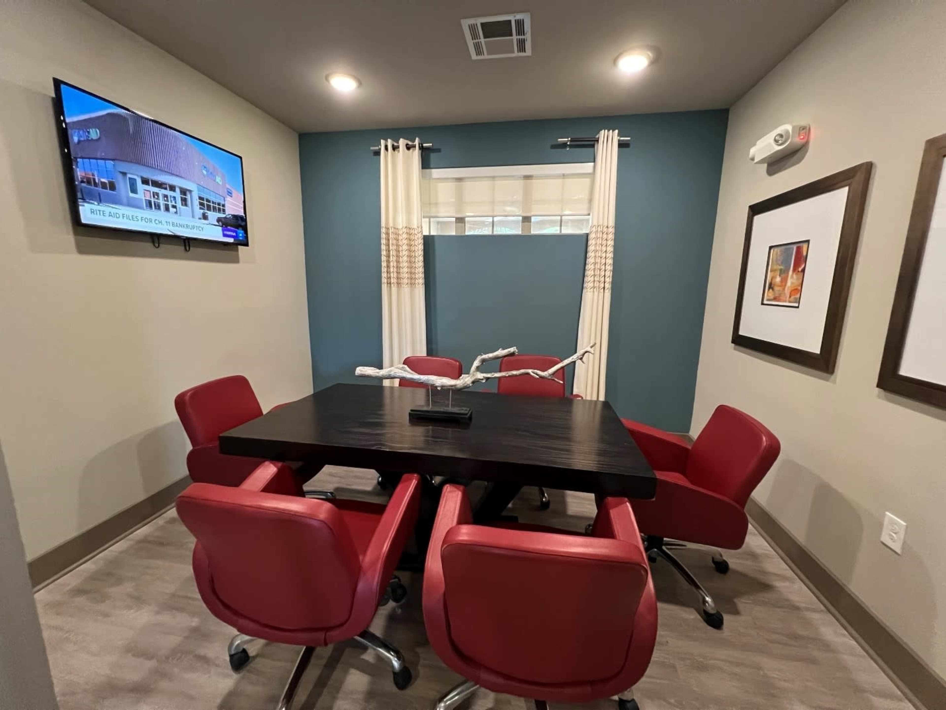 The image shows a small conference room with a dark wooden table surrounded by four red chairs, a television on the wall, and large windows dressed with curtains.