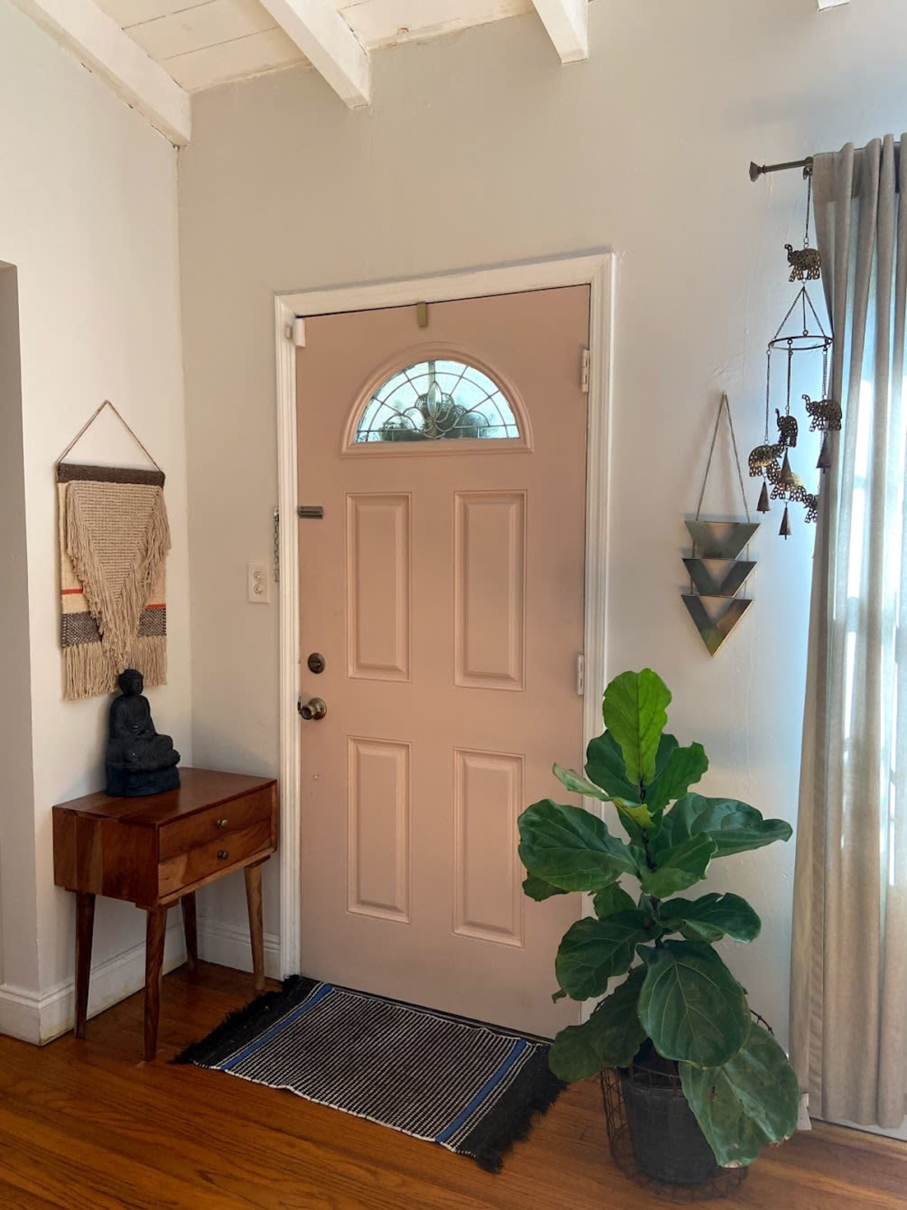 A light-colored entryway featuring a pink front door, a small wooden table with a statue, a potted plant, and a window with sheer curtains.