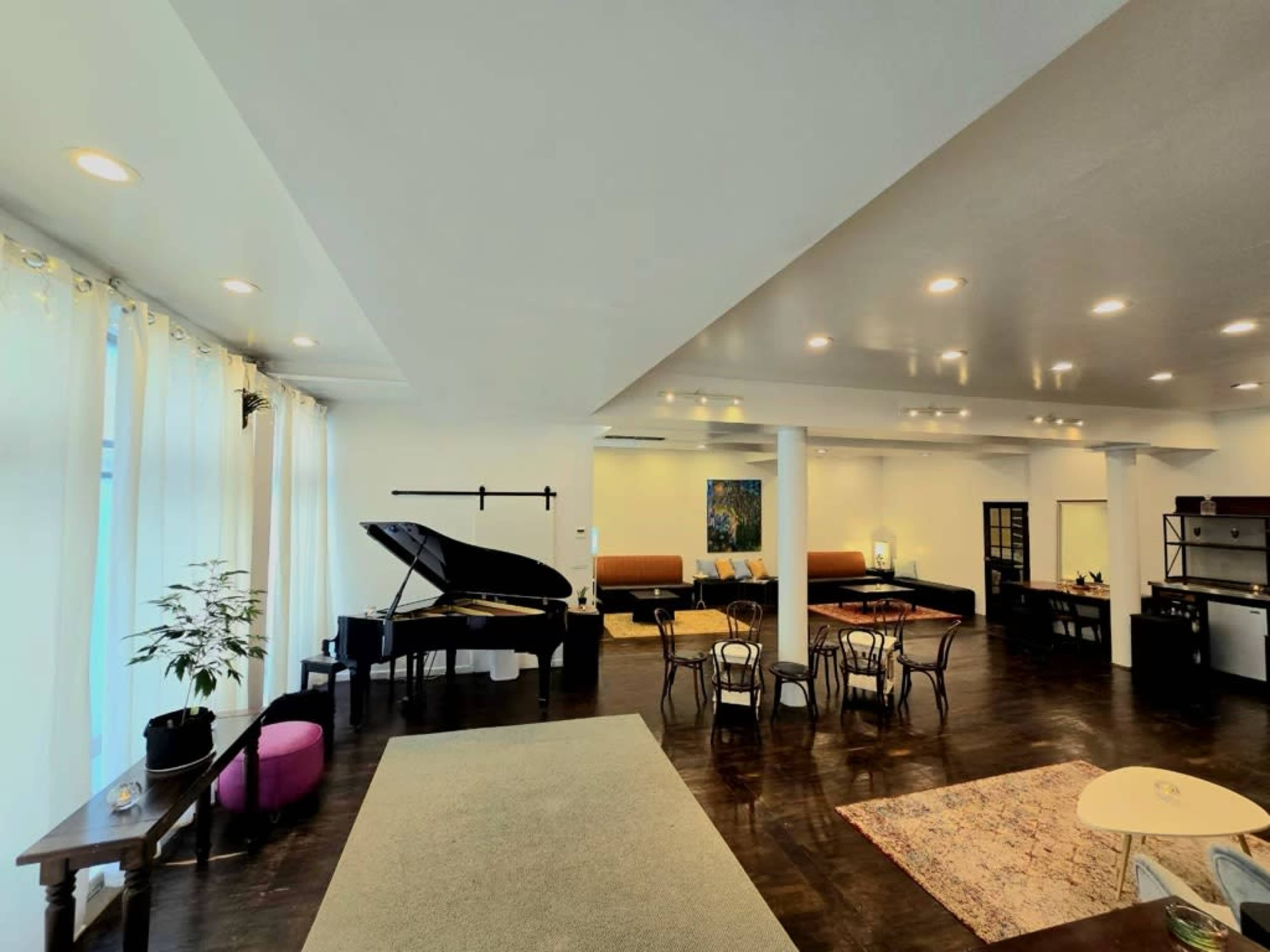 The image shows a spacious room featuring a grand piano, several dining chairs around a table, and a modern kitchen area.