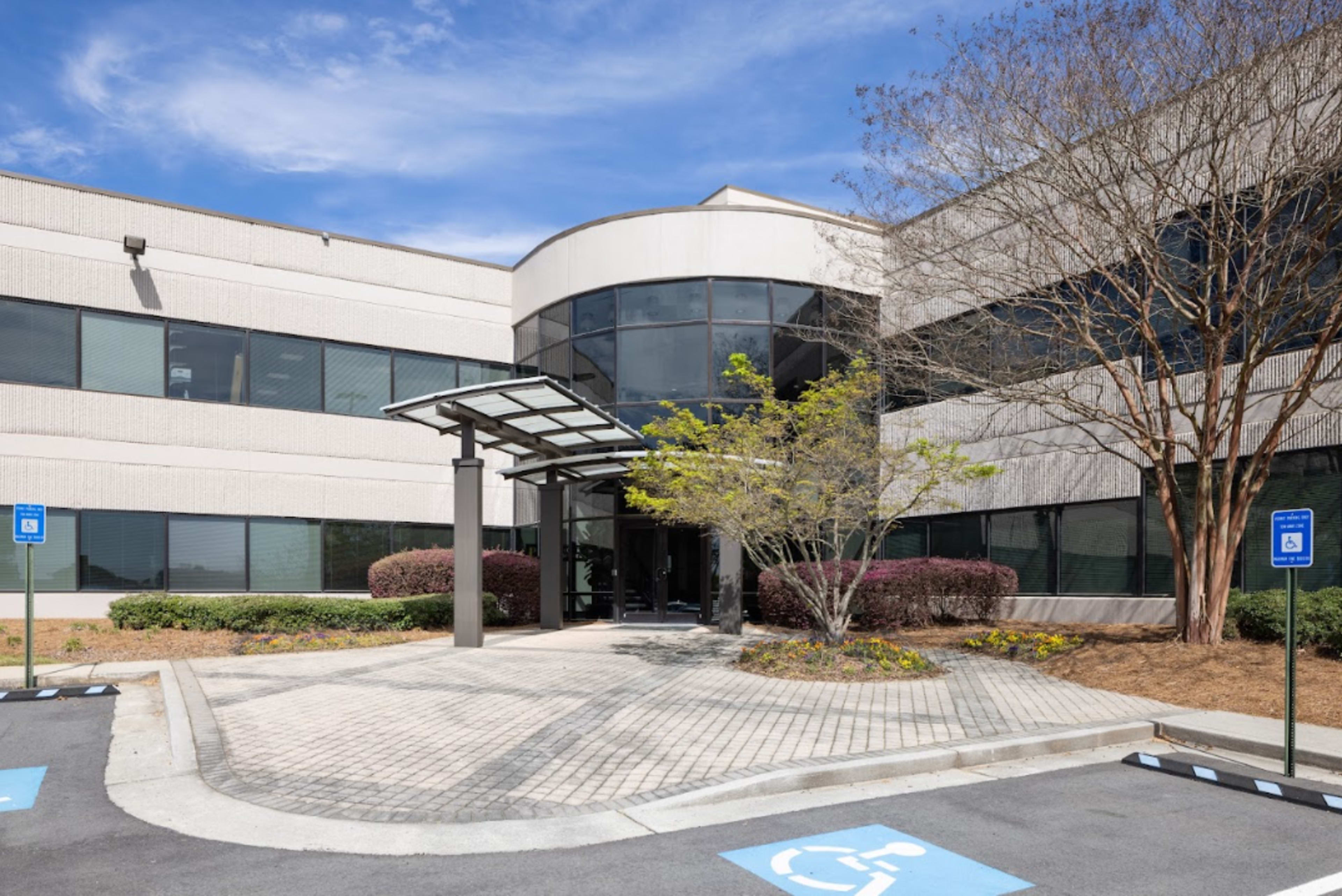 The image shows the exterior of a modern office building with large windows, a covered entrance, and landscaped surroundings.