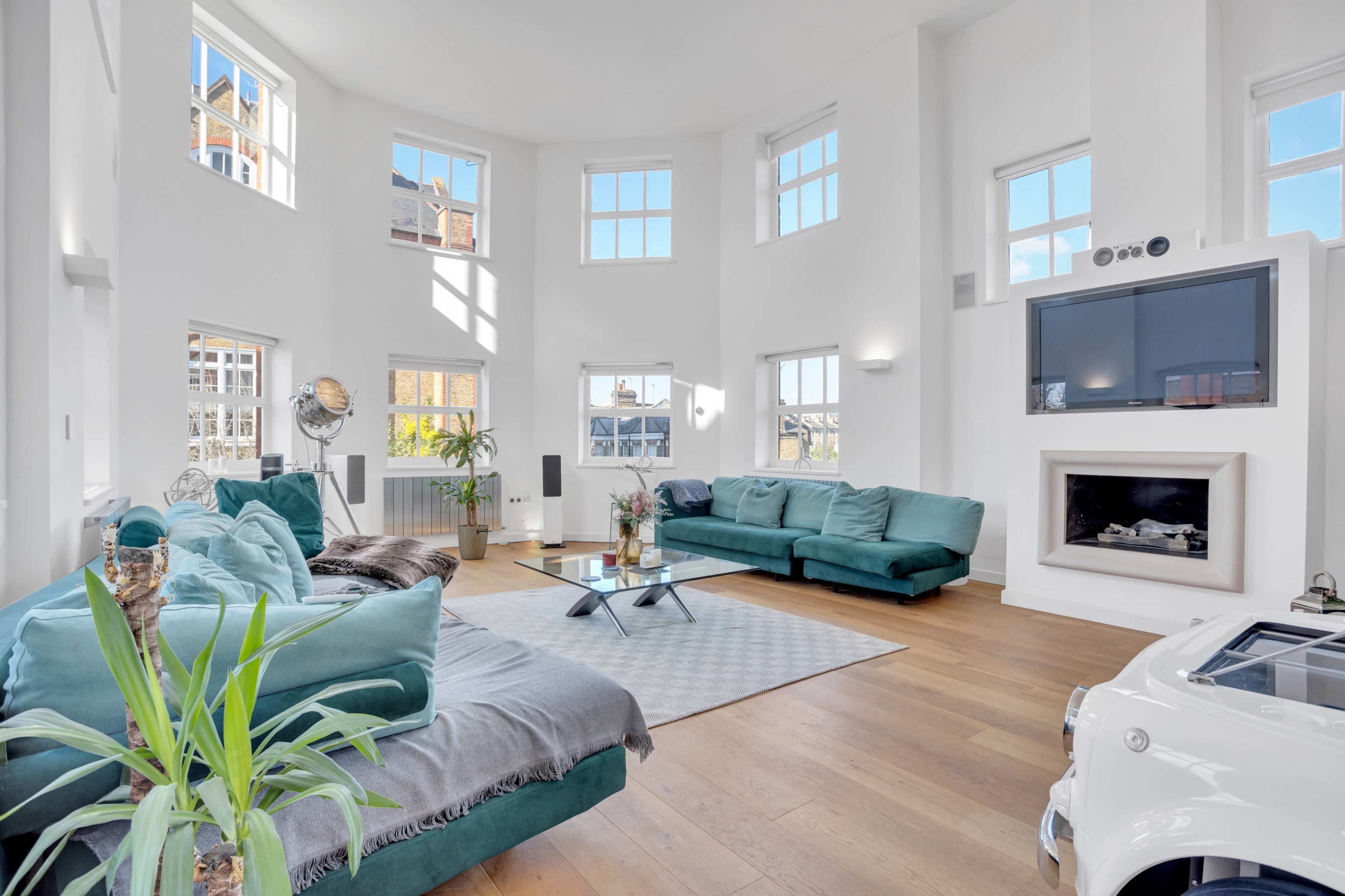 The image shows a bright, modern living room featuring two green sofas, large windows, and a fireplace.