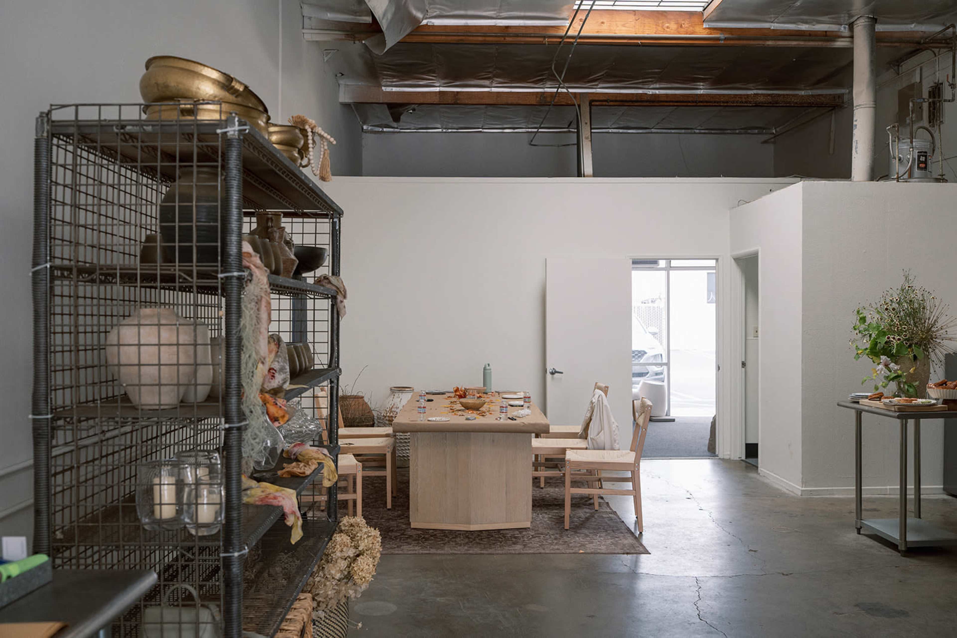 The image shows a minimalistic interior space featuring a dining table set for a meal, a storage shelf with various items, and a white door leading outside.