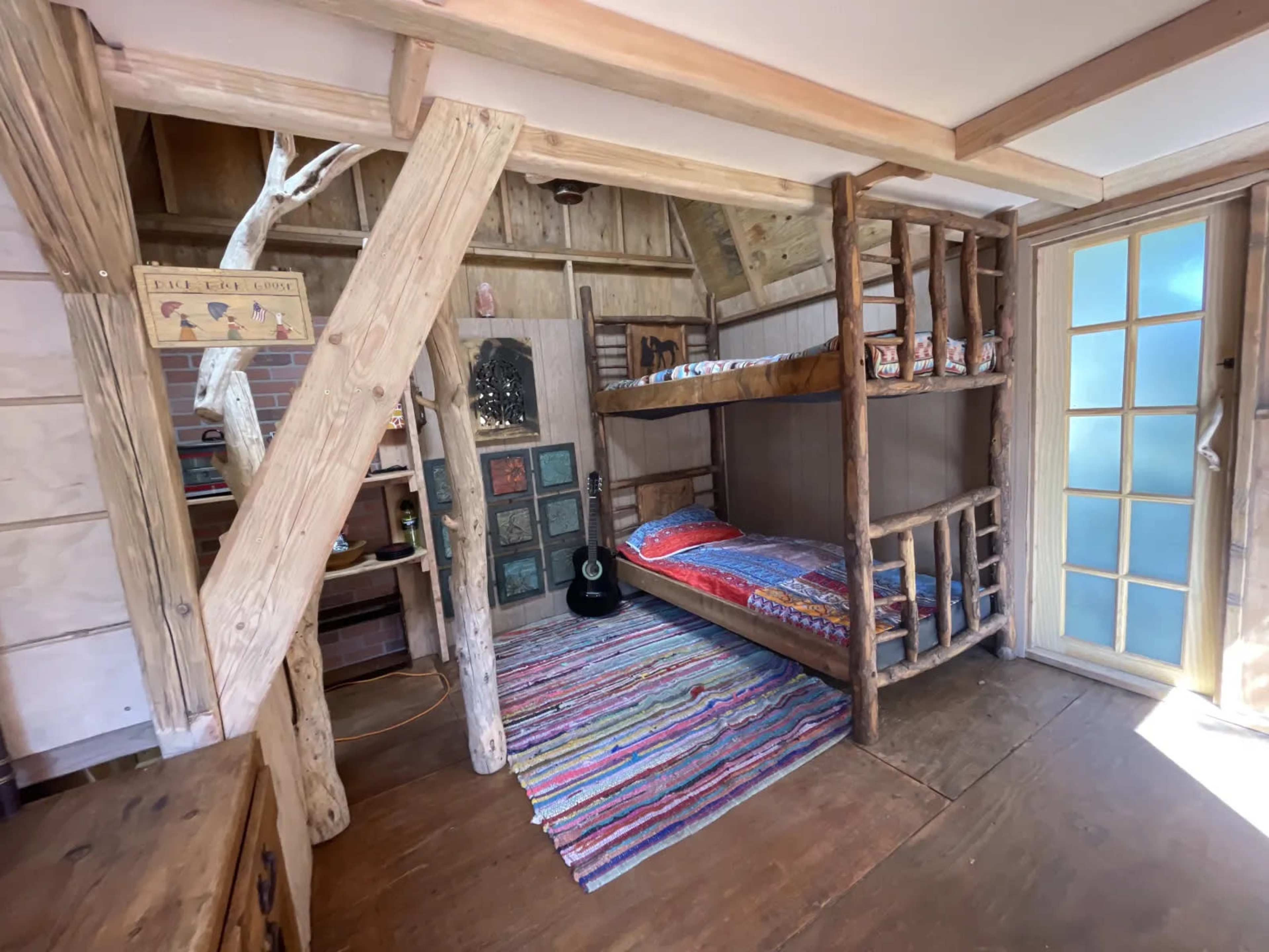The image shows a rustic bedroom featuring a bunk bed made of wooden logs, a colorful woven rug on the floor, and a guitar leaning against the wall.