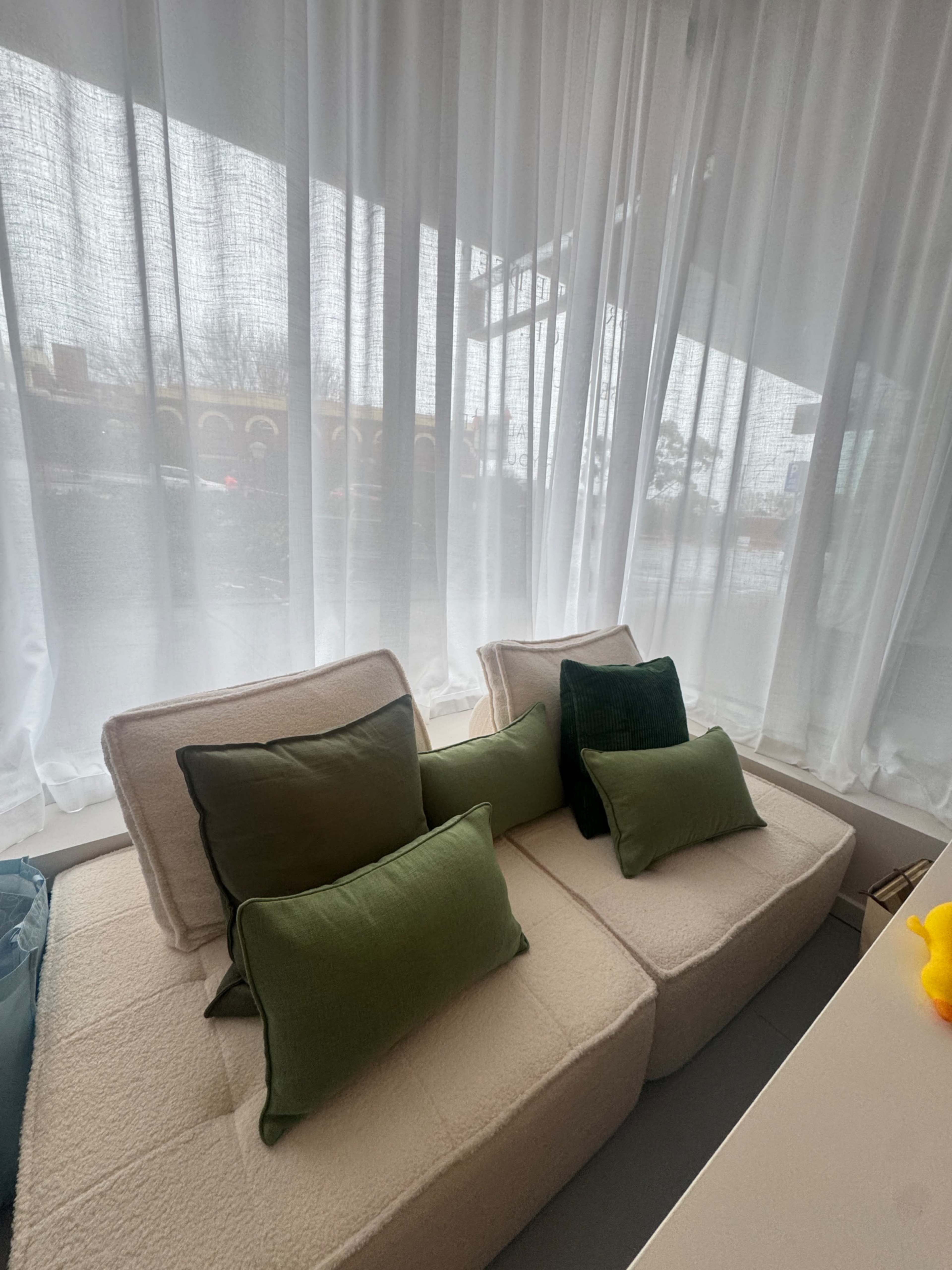 A cozy seating area features cushions in various shades of green against a backdrop of sheer white curtains and a window showing a gray, rainy day outside.