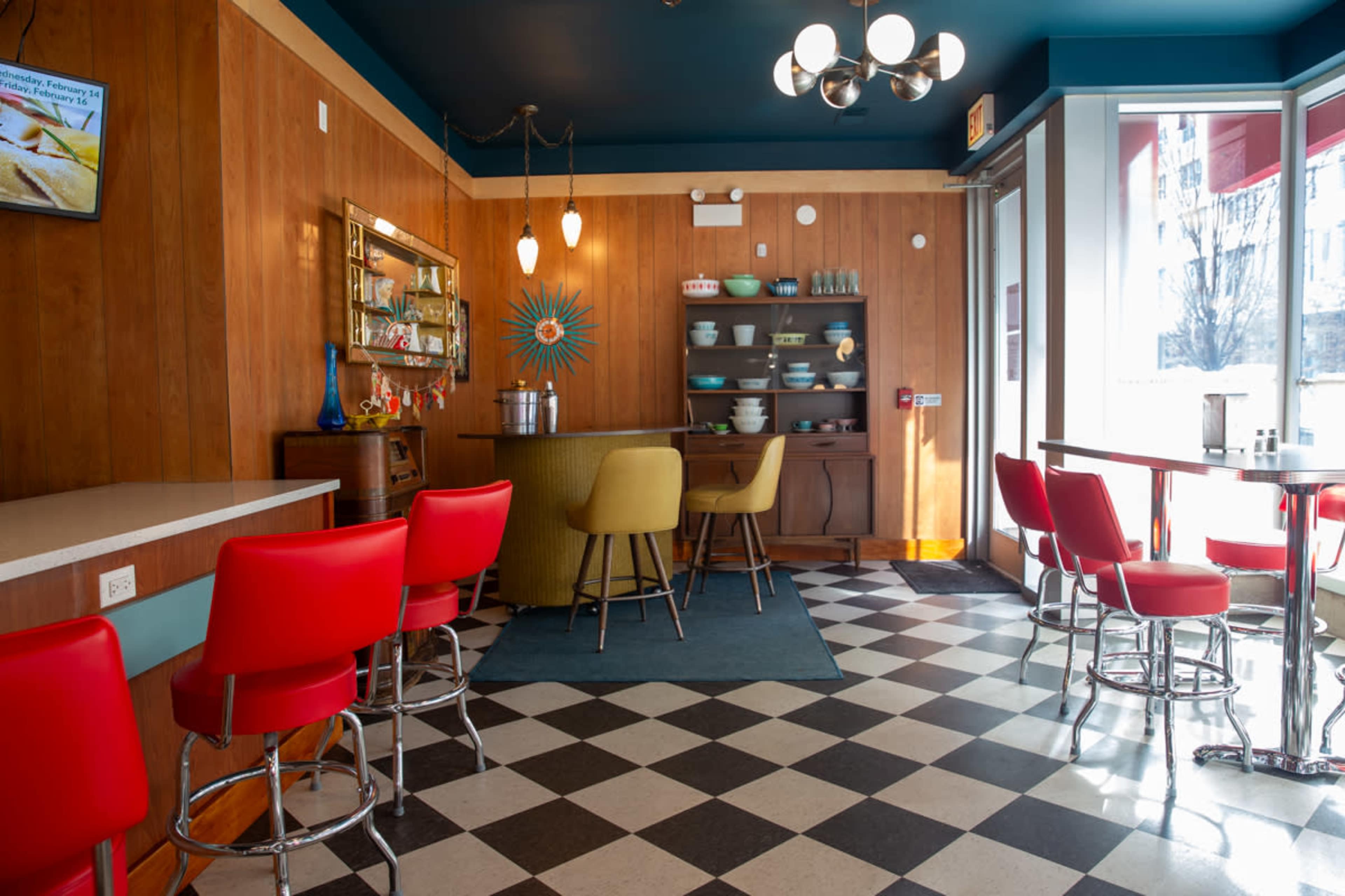 A colorful cafe features a checkered floor, retro seating, and a decorative bar area with various glassware and dishware on display.