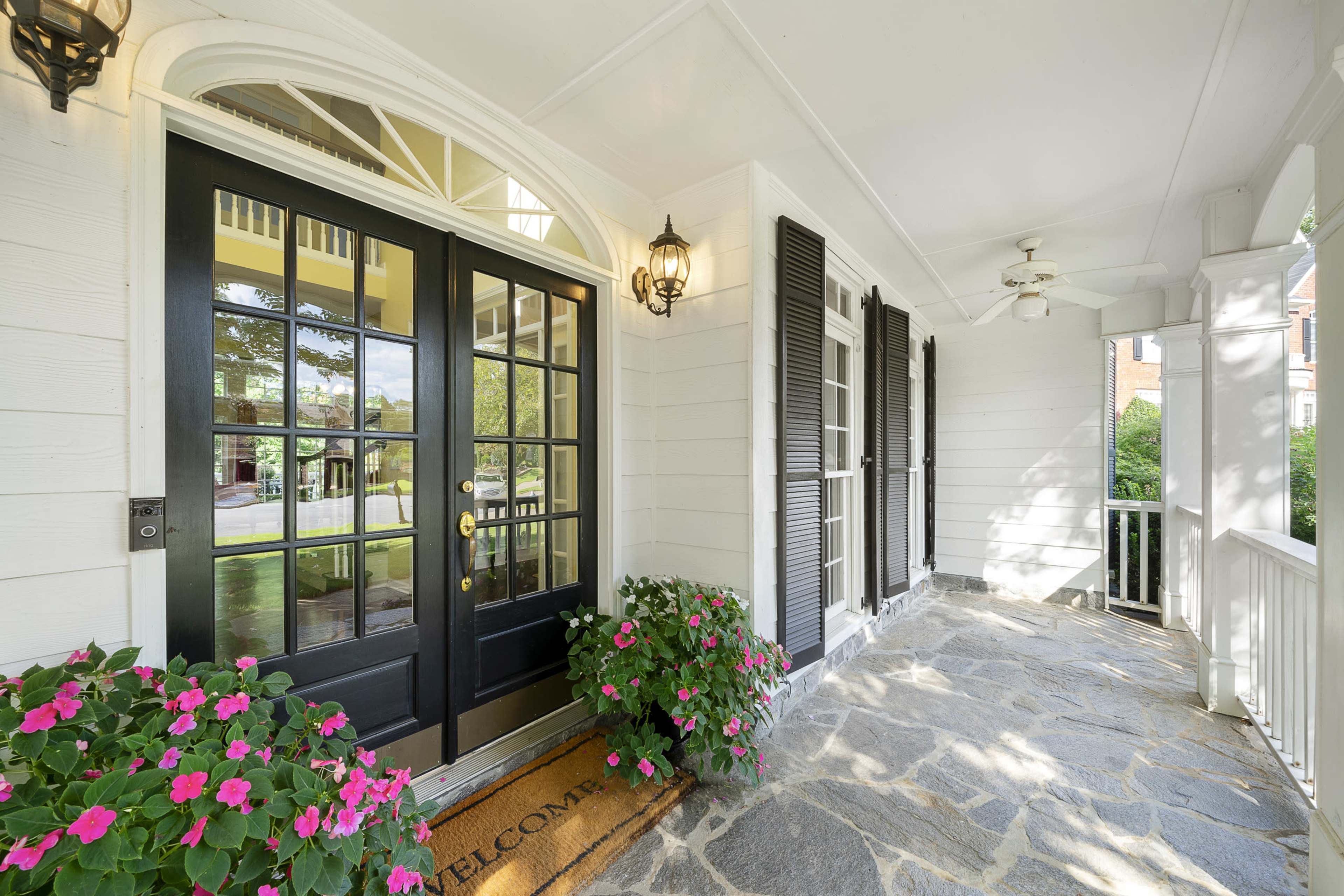 A covered porch with two black doors, a welcome mat, potted flowers, and ceiling fans.
