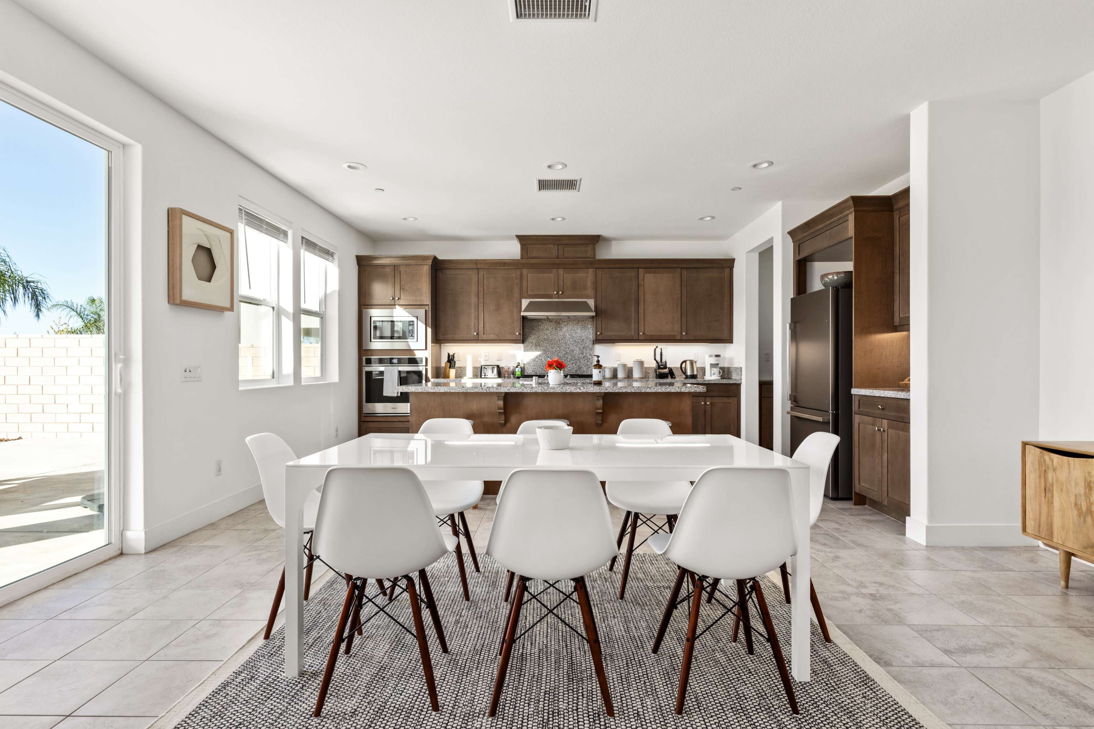 A modern kitchen features dark wooden cabinets, stainless steel appliances, and a dining table surrounded by white chairs on a patterned rug.