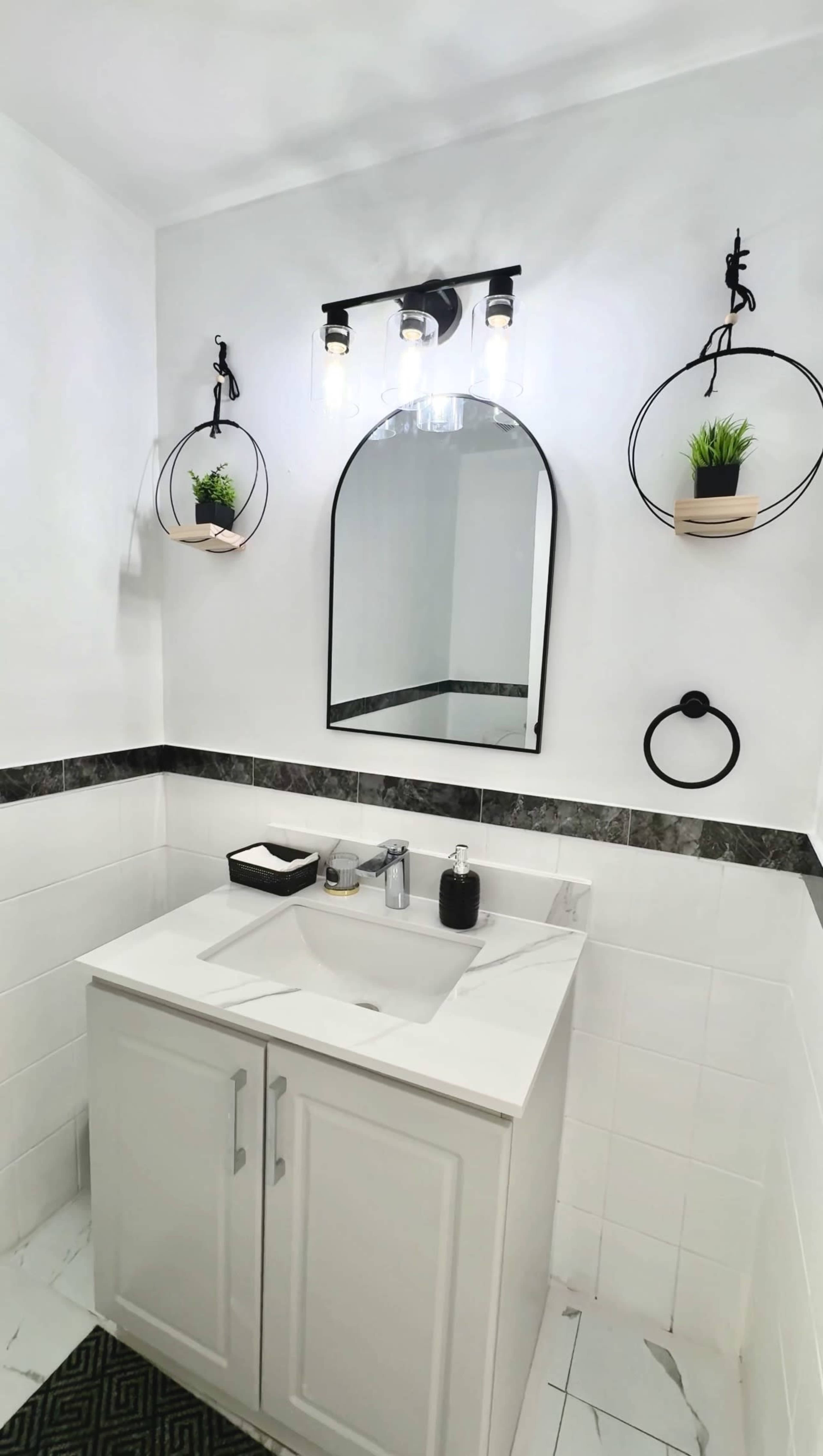 The image shows a modern bathroom with a white vanity, a round mirror above, and decorative plants in circular wall-mounted holders.