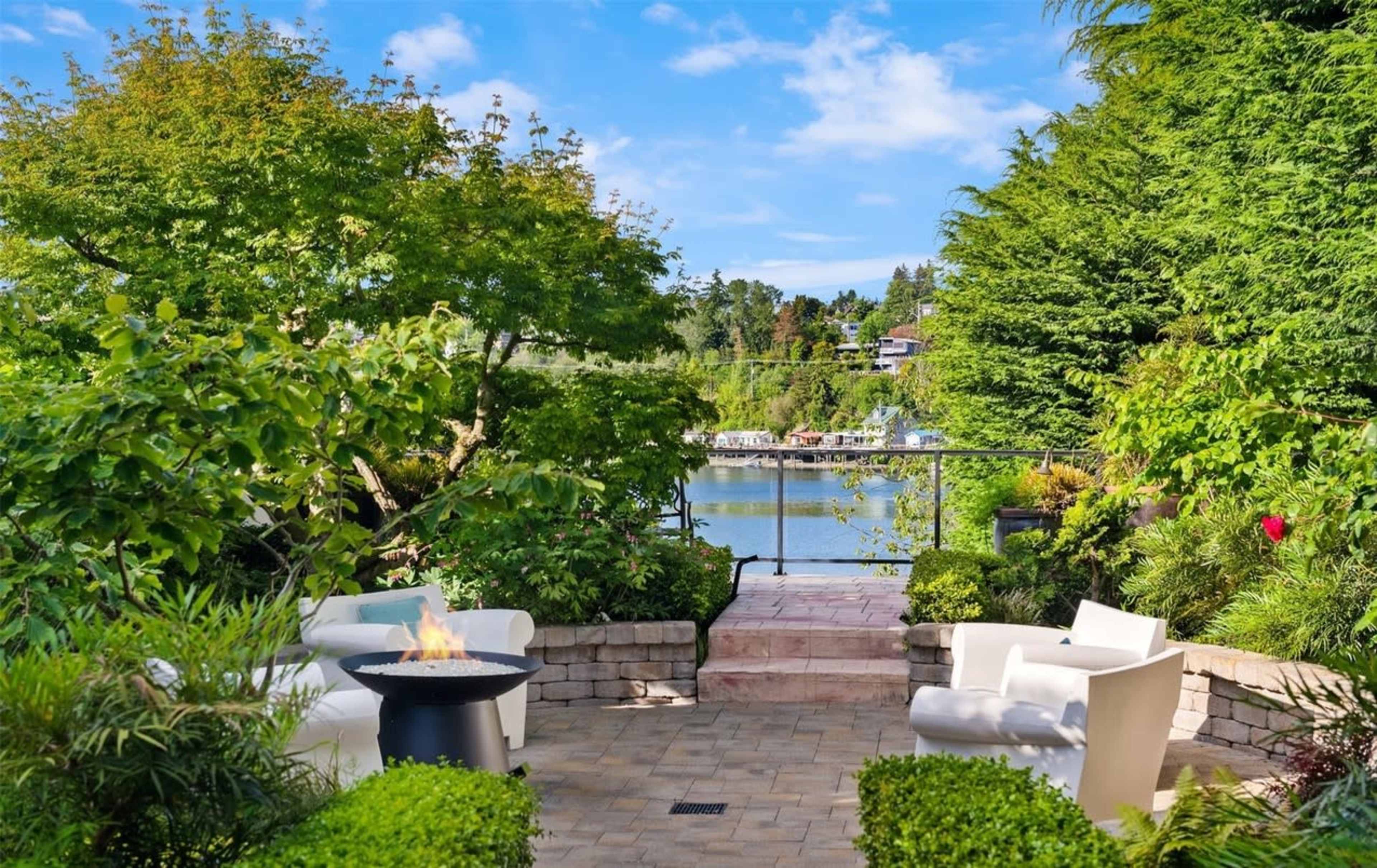 A landscaped outdoor area features white seating and a fire pit overlooking a tranquil body of water surrounded by greenery.