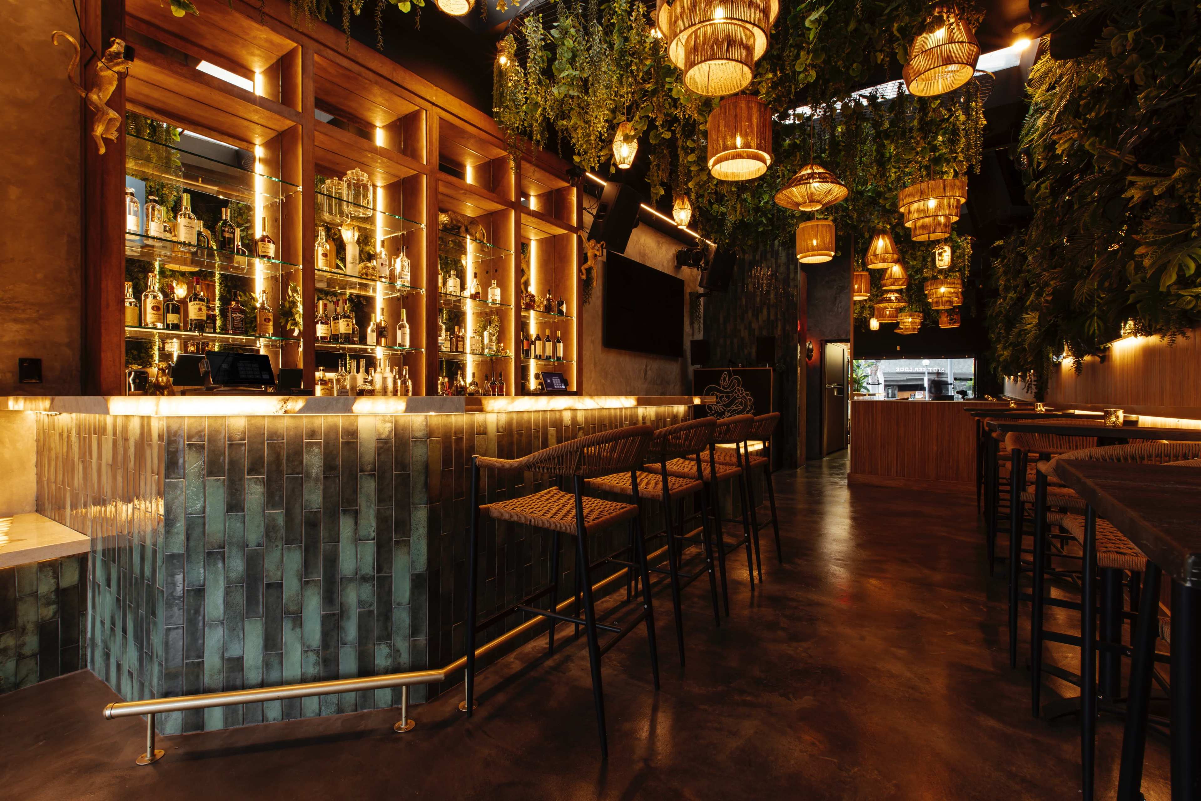 A stylish bar interior featuring a well-stocked display behind the bar, surrounded by hanging plants and lanterns, with high stools arranged in front.