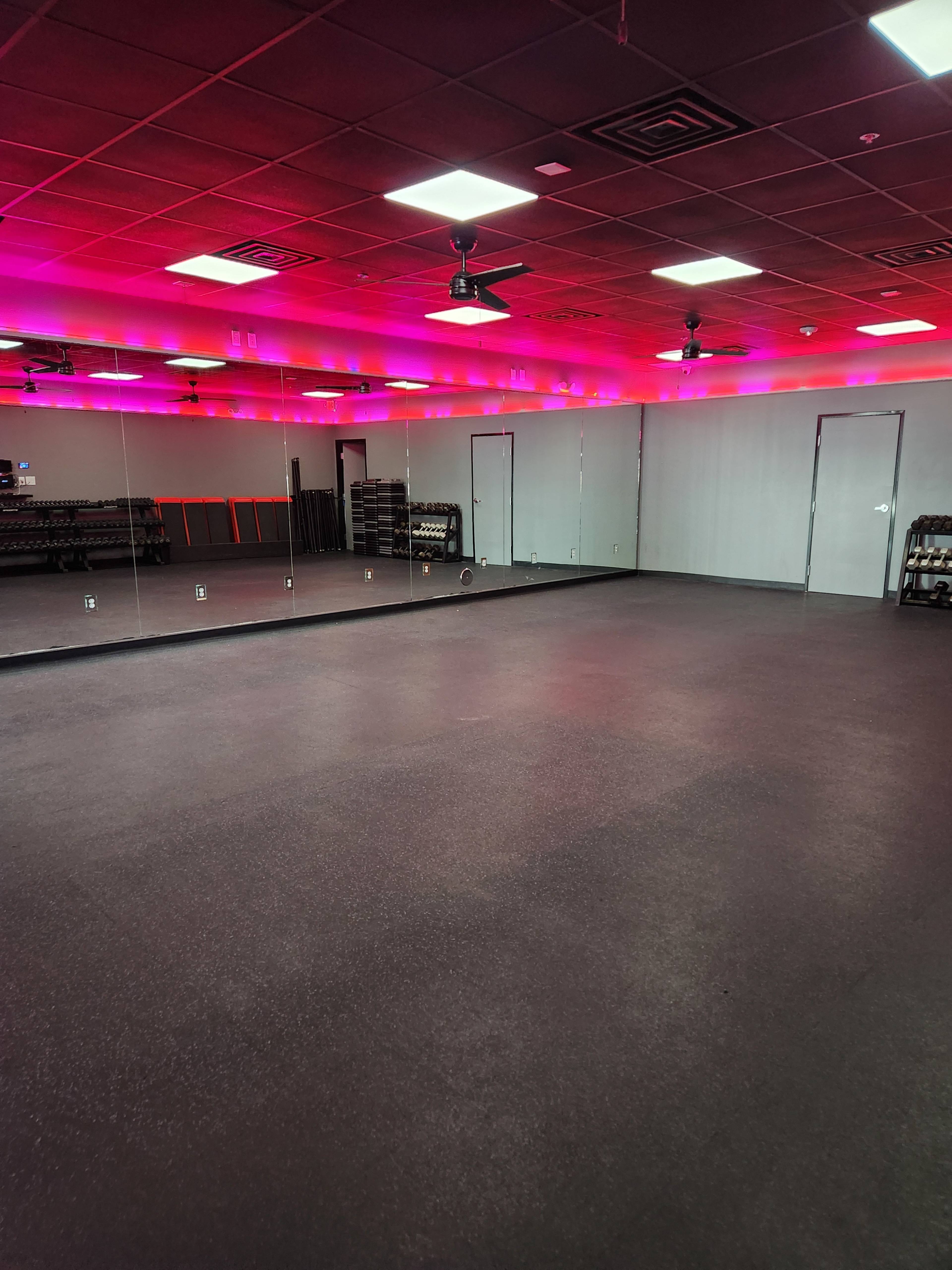 An exercise studio with a mirrored wall, dark flooring, and lighting that creates a pink hue along the ceiling.