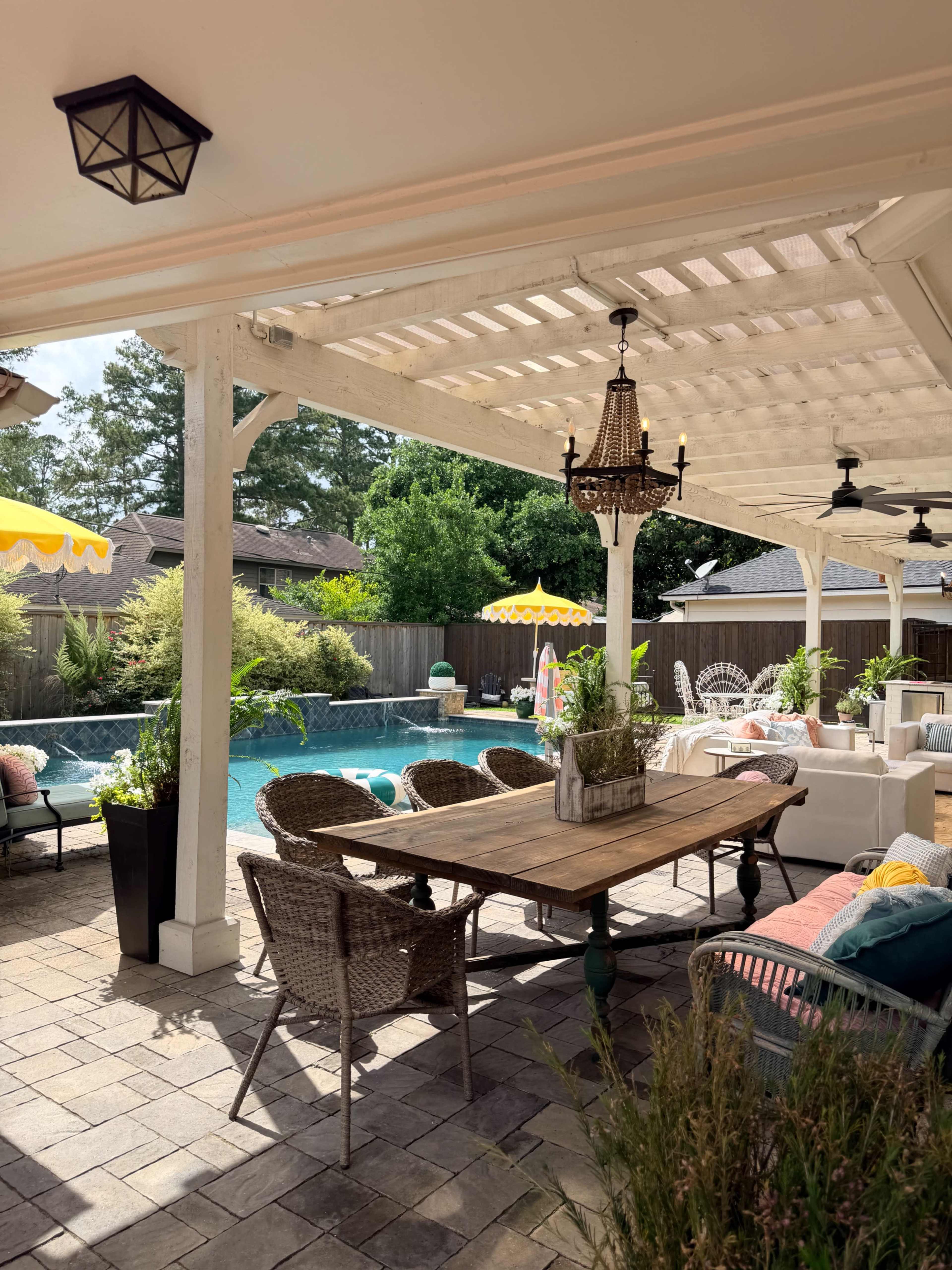 The image shows a covered outdoor patio area featuring a wooden dining table with wicker chairs, a swimming pool in the background, and yellow umbrellas providing shade.