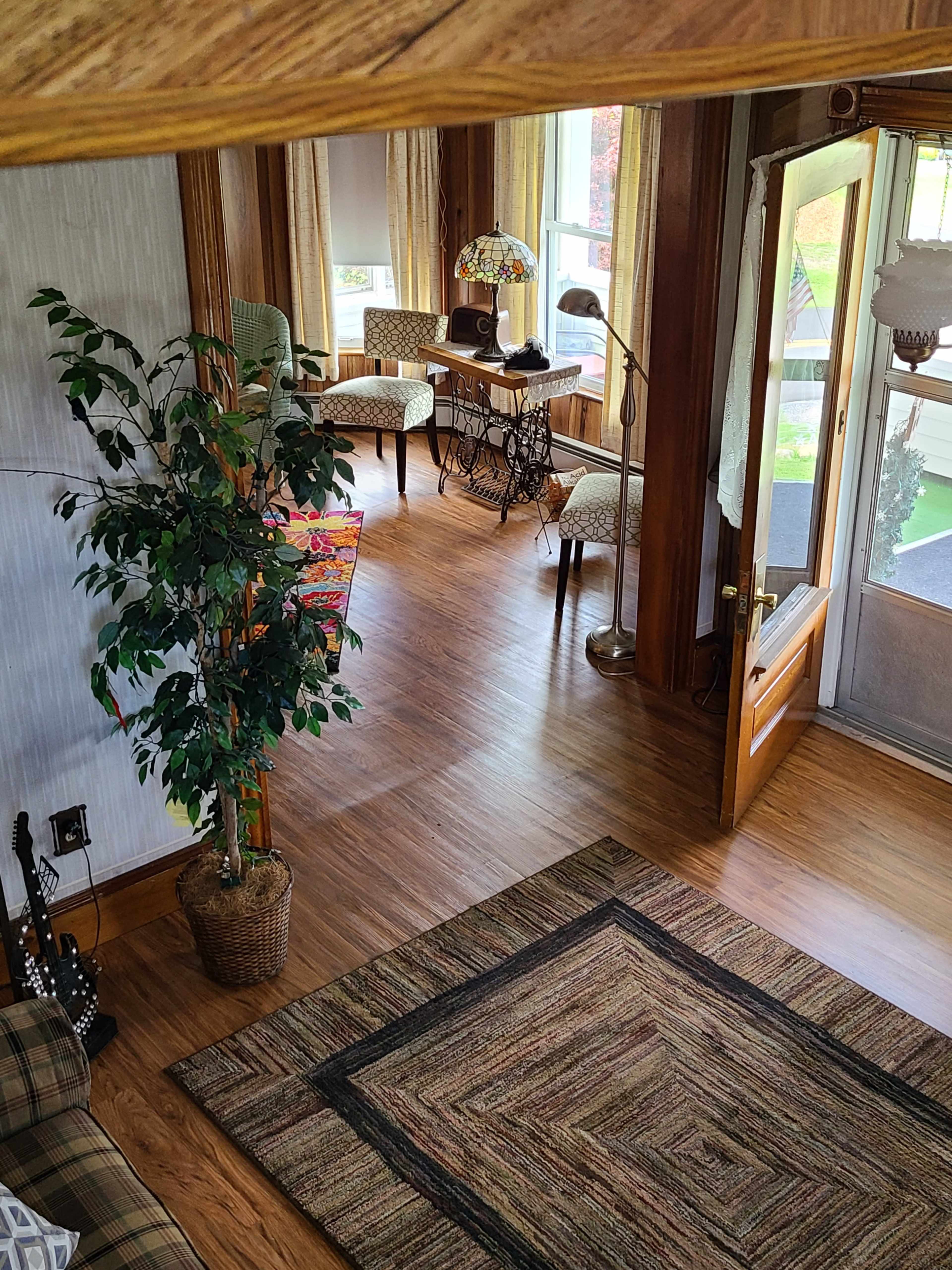 A cozy living room with wooden beams, a patterned rug, a potted plant, and a seating area that includes a table and chairs beside a large window.